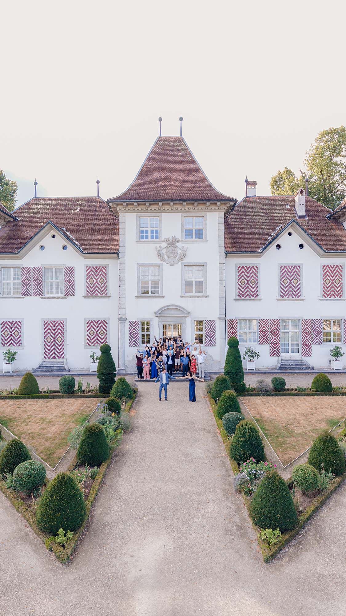 Aerial view of elegant French château with geometric topiary gardens and wedding guests gathered at the entrance.