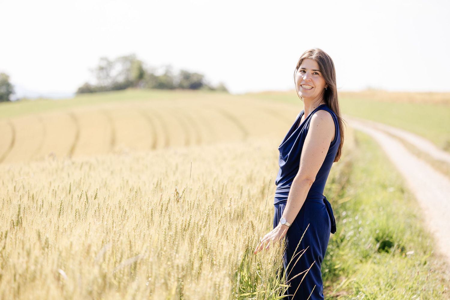 Woman in navy blue shirt poses in sunny wheat field during golden hour with hair blowing in the breeze.
