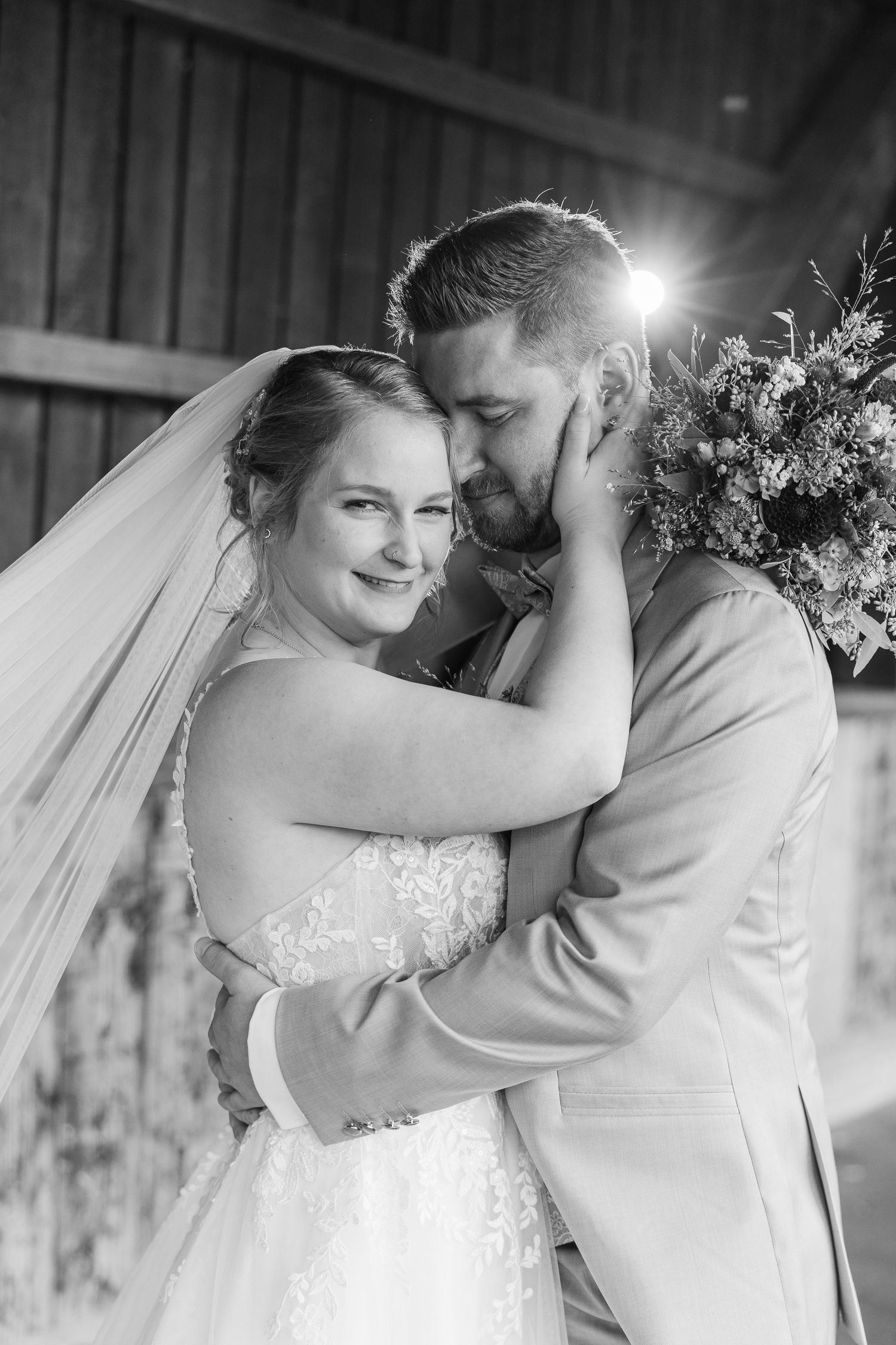 Black and white photo of a smiling bride in a lace gown and veil embracing her groom, who holds a bouquet, in a rustic barn.