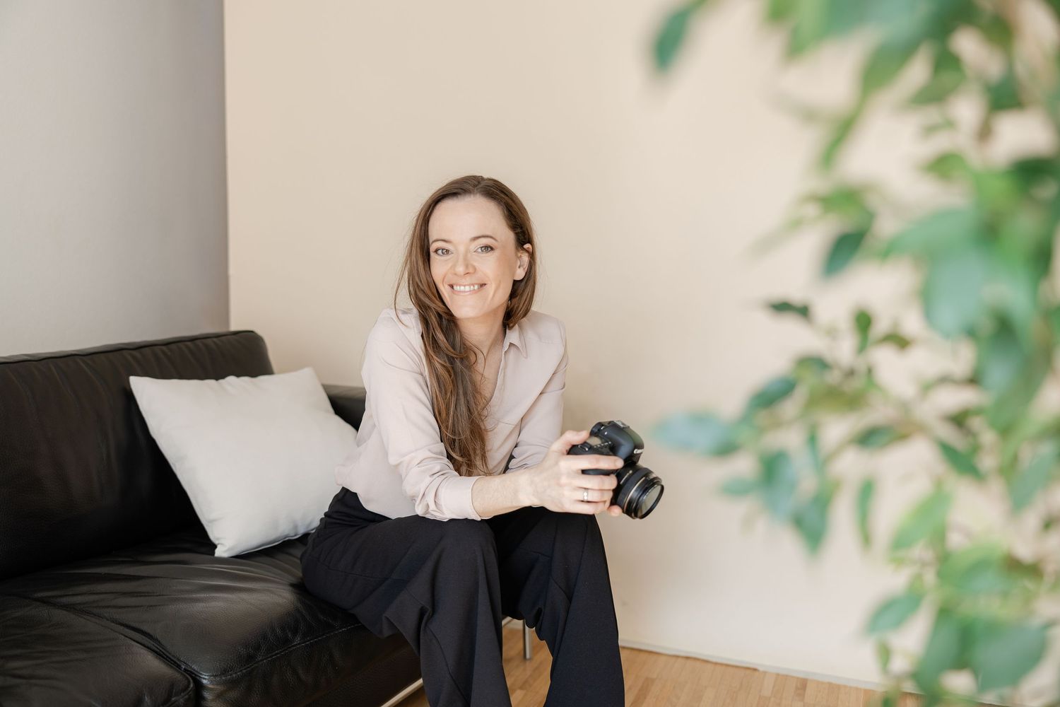 A photographer with a camera sits casually on a black couch near indoor plants and white pillows. Aarau, Schweiz