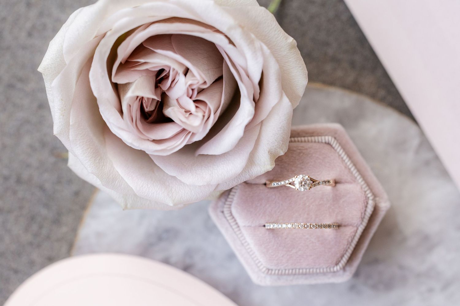 Pink rose beside a velvet hexagon ring box with delicate gold diamond engagement ring and bracelet on marble surface.