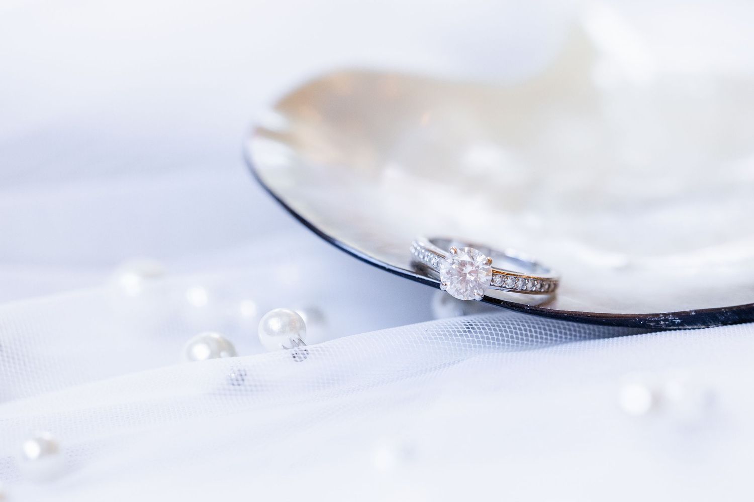 Diamond engagement ring with pavé band resting on white pearl shell, surrounded by scattered pearls on soft white fabric.