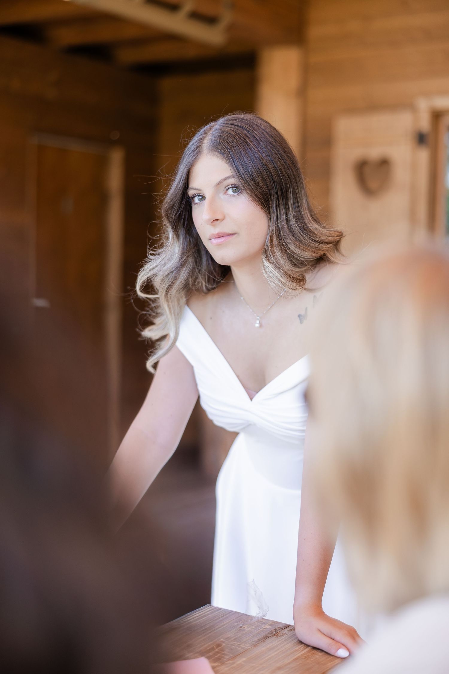 Bride in white off-shoulder gown standing in rustic wooden venue, looking elegantly over her shoulder.