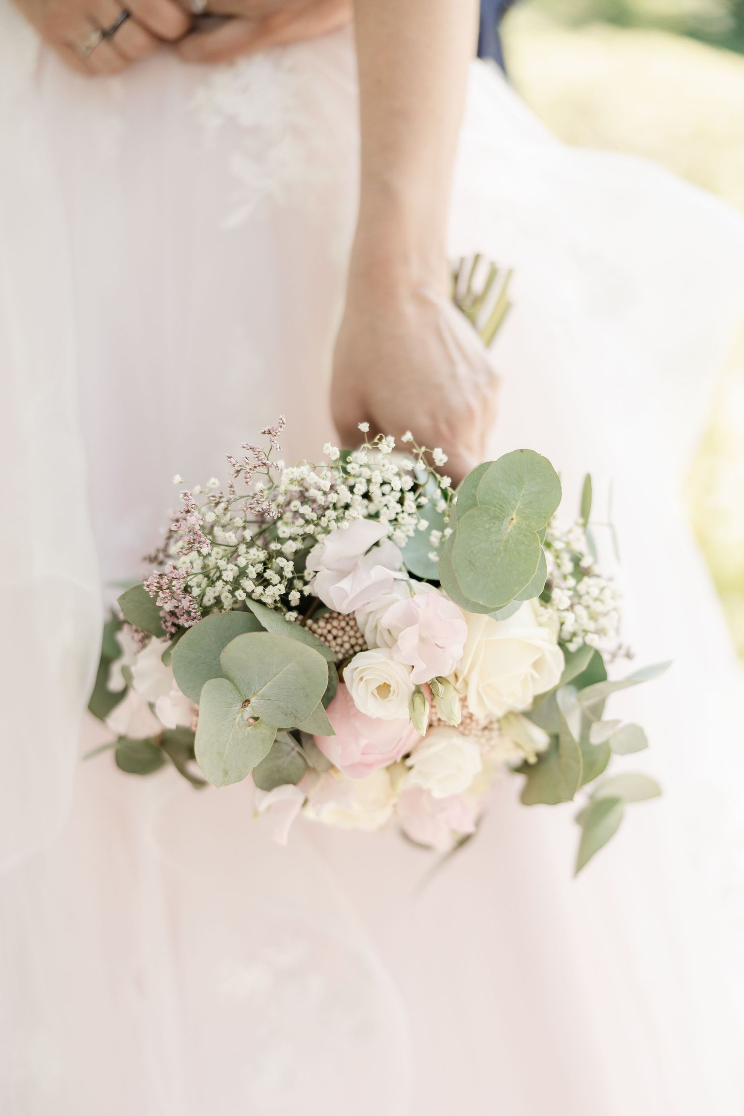 A delicate bridal bouquet featuring white and blush roses with eucalyptus leaves and baby's breath against a white dress.
