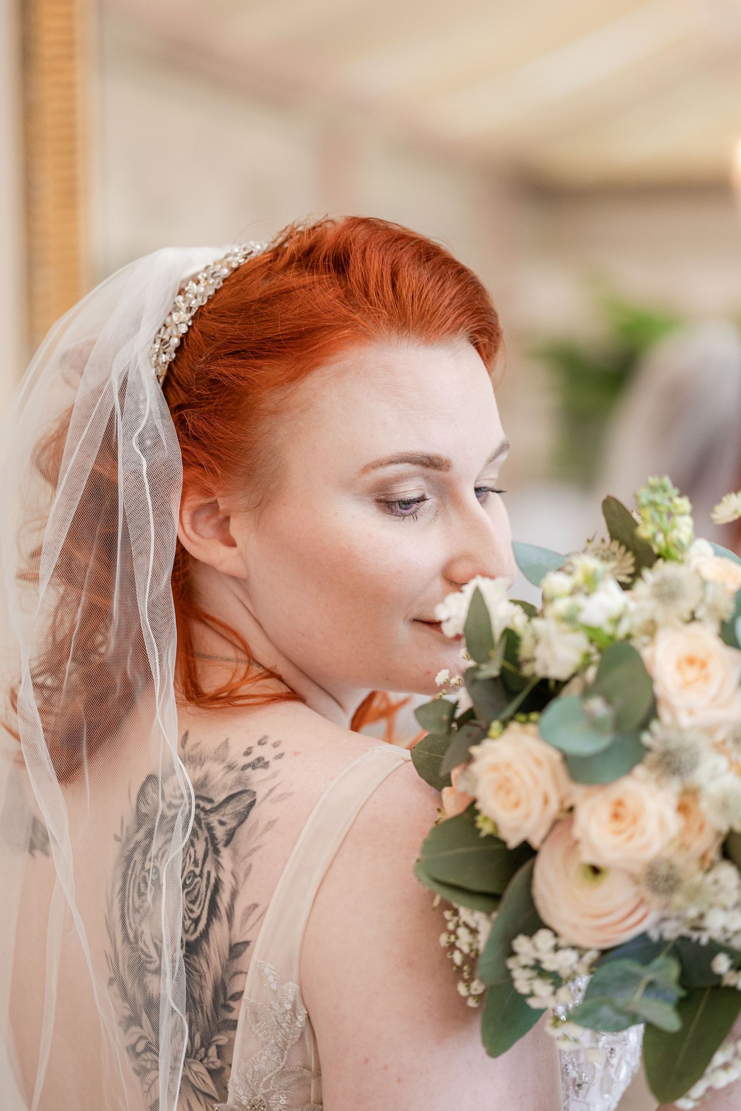 Red-haired bride in veil and lace dress smelling white and peach rose bouquet, with visible back tattoo.