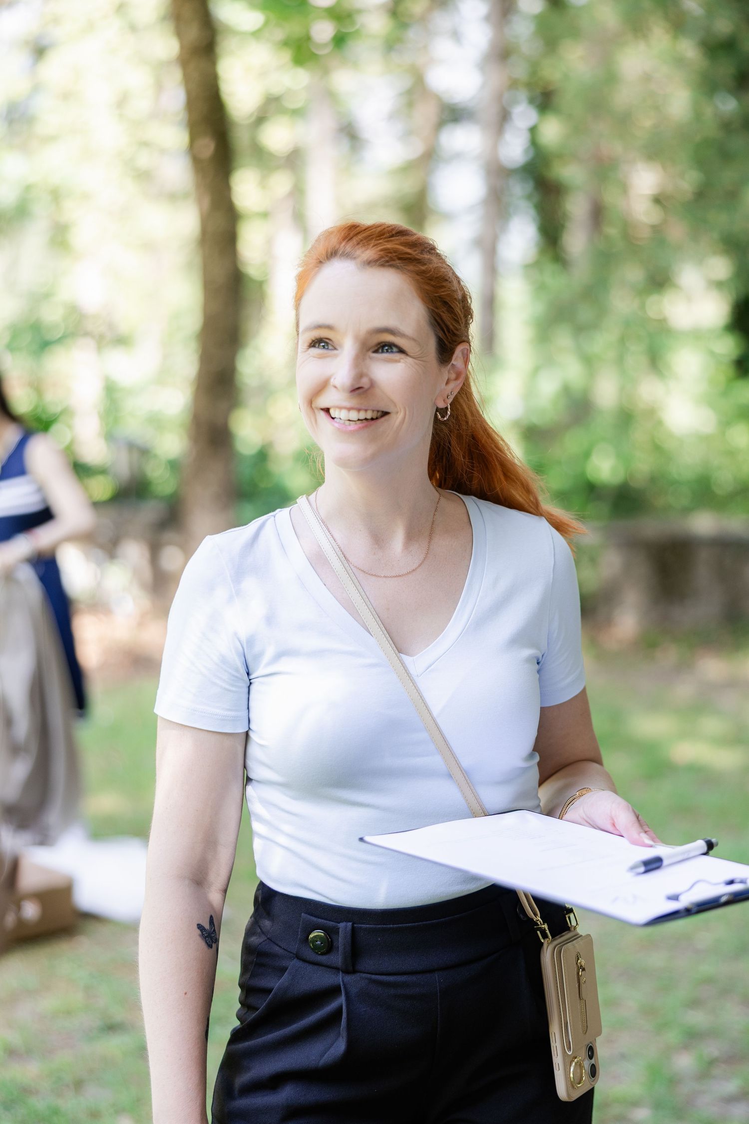 Smiling redhead woman holding clipboard outdoors in green park, wearing white t-shirt and black pants with crossbody bag.