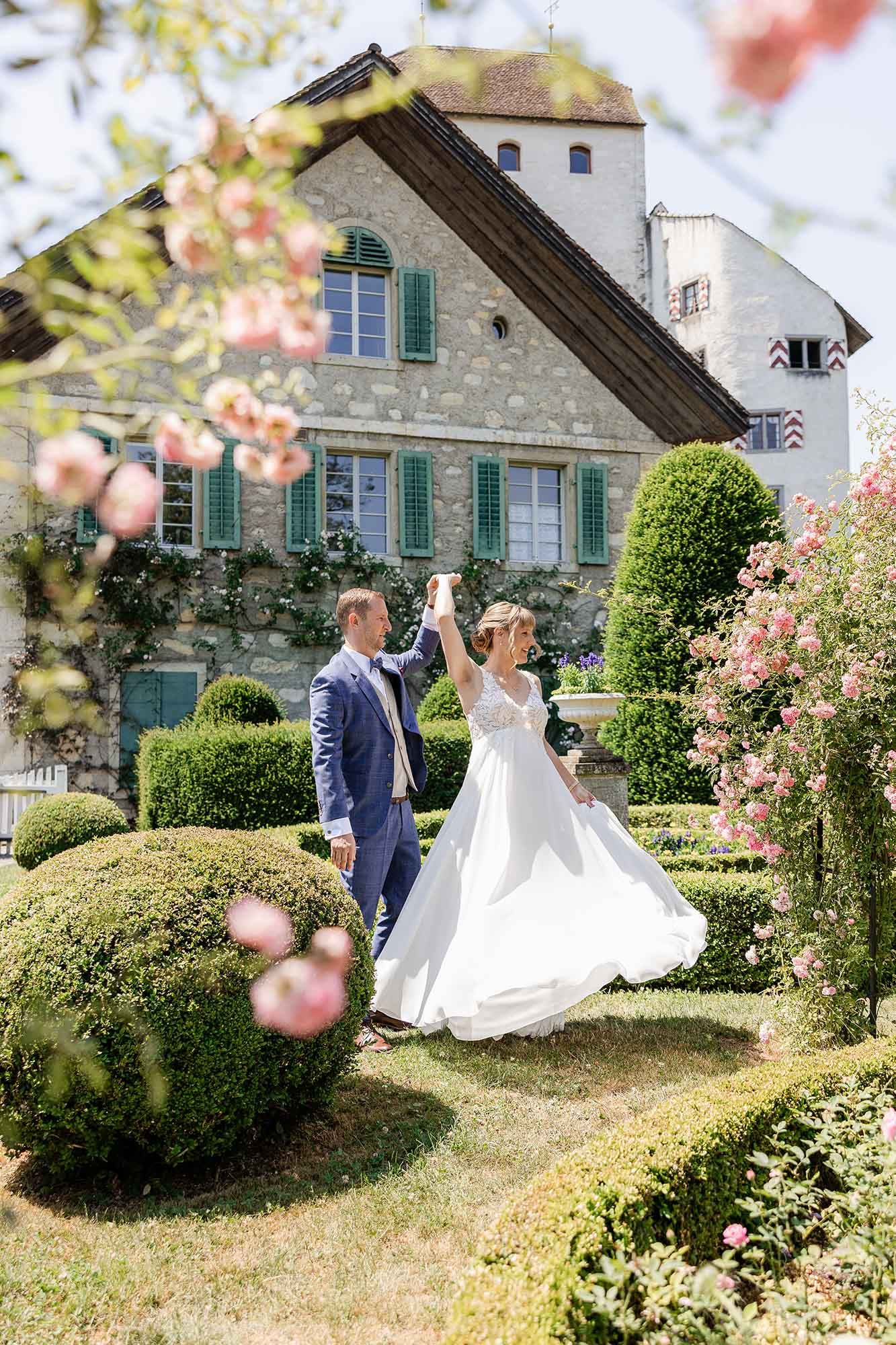 Bride and groom dancing in a romantic garden with blooming roses and manicured hedges in front of a charming stone manor.