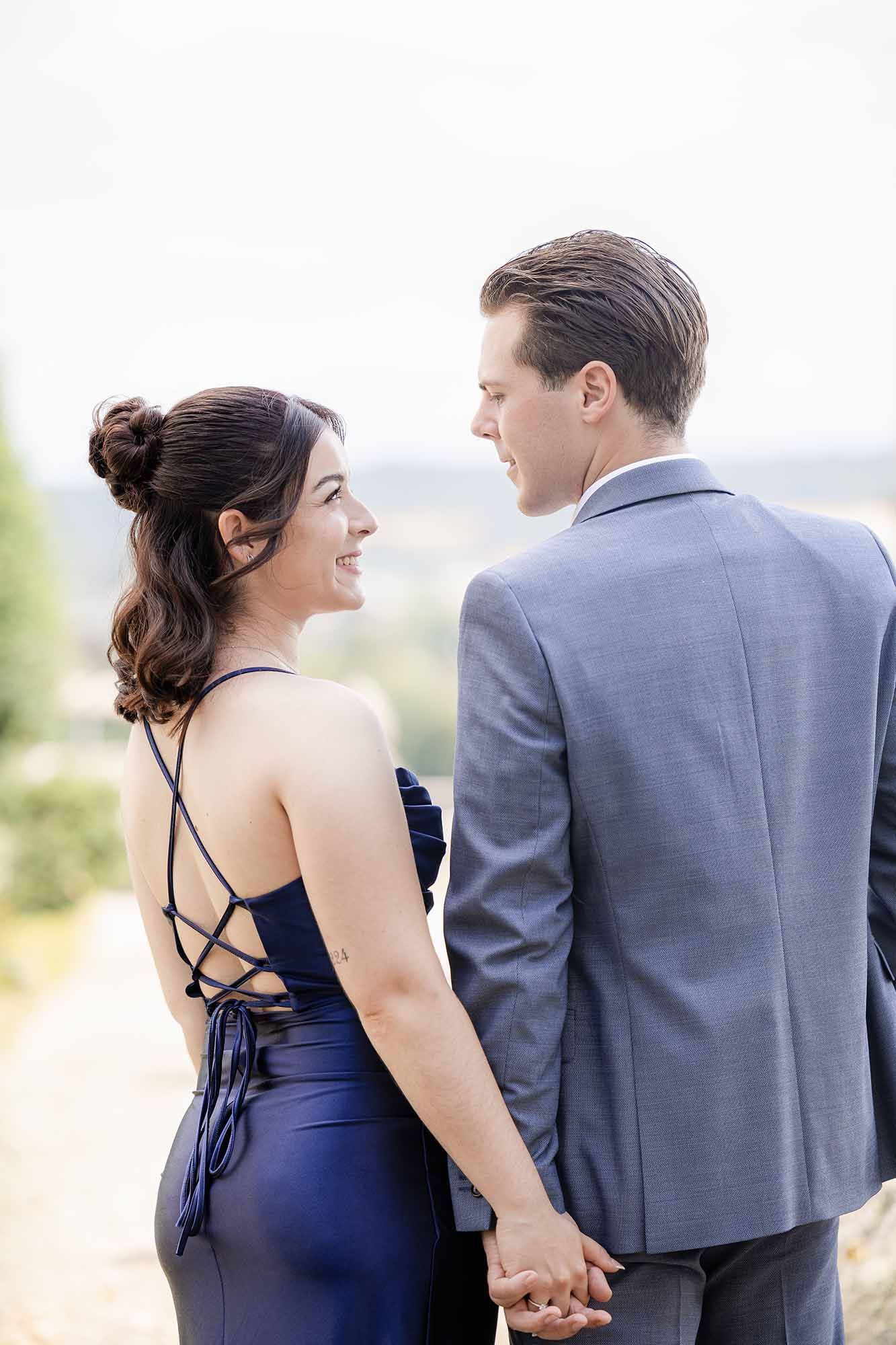 Couple holding hands in formal wear, woman in navy blue backless dress smiling at man in gray suit outdoors.