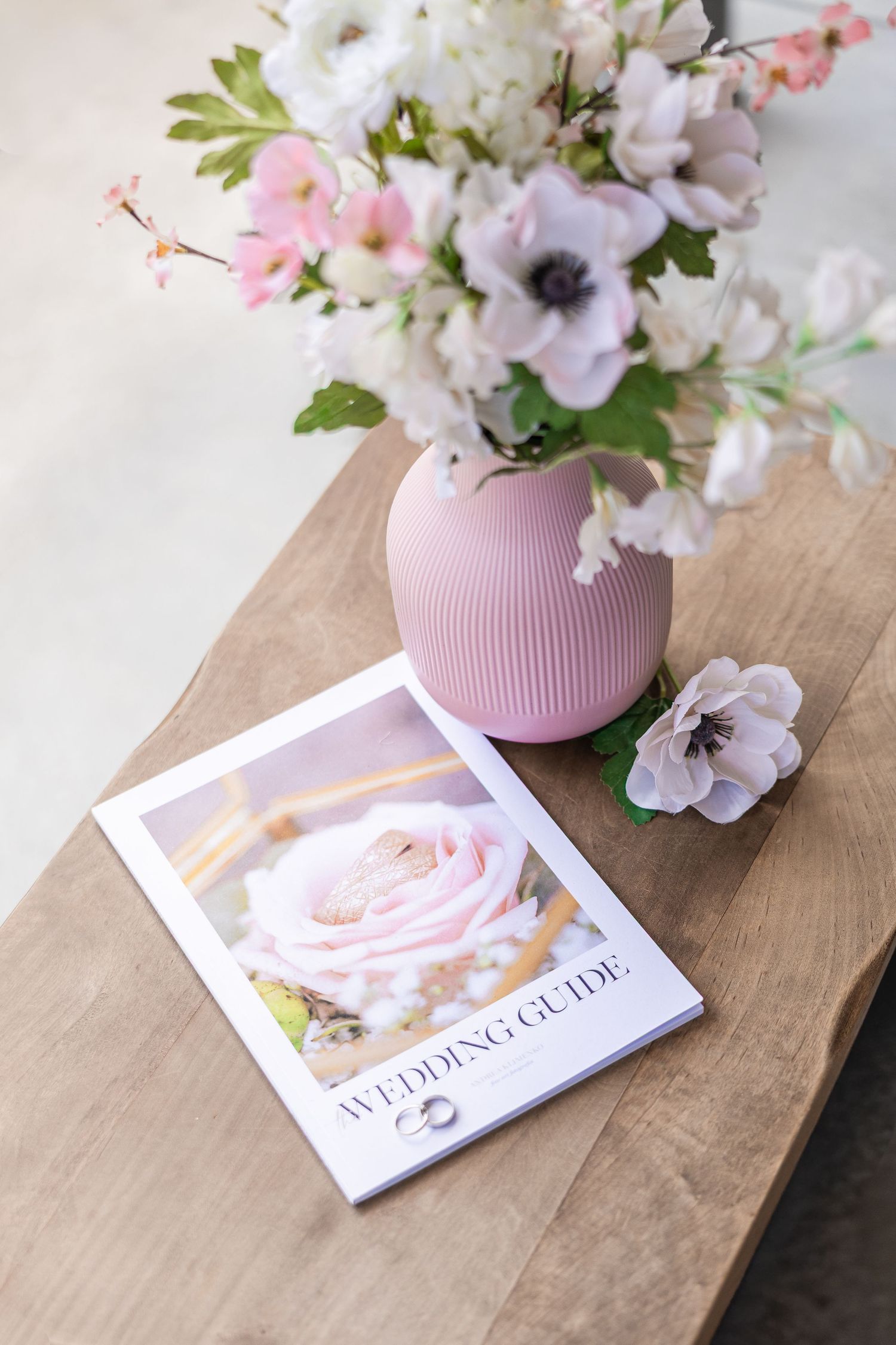 A wedding guide booklet beside a pink vase with anemone flowers on a wooden table.