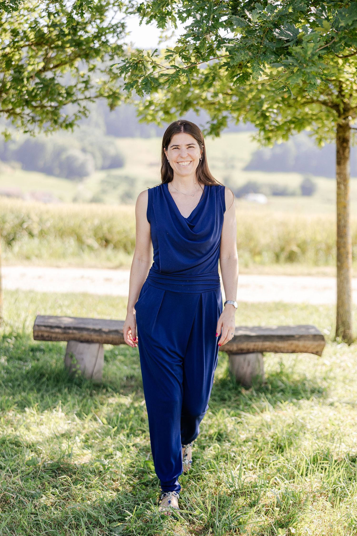 A person in a navy blue dress poses outdoors in a sunlit field with trees in the background.
