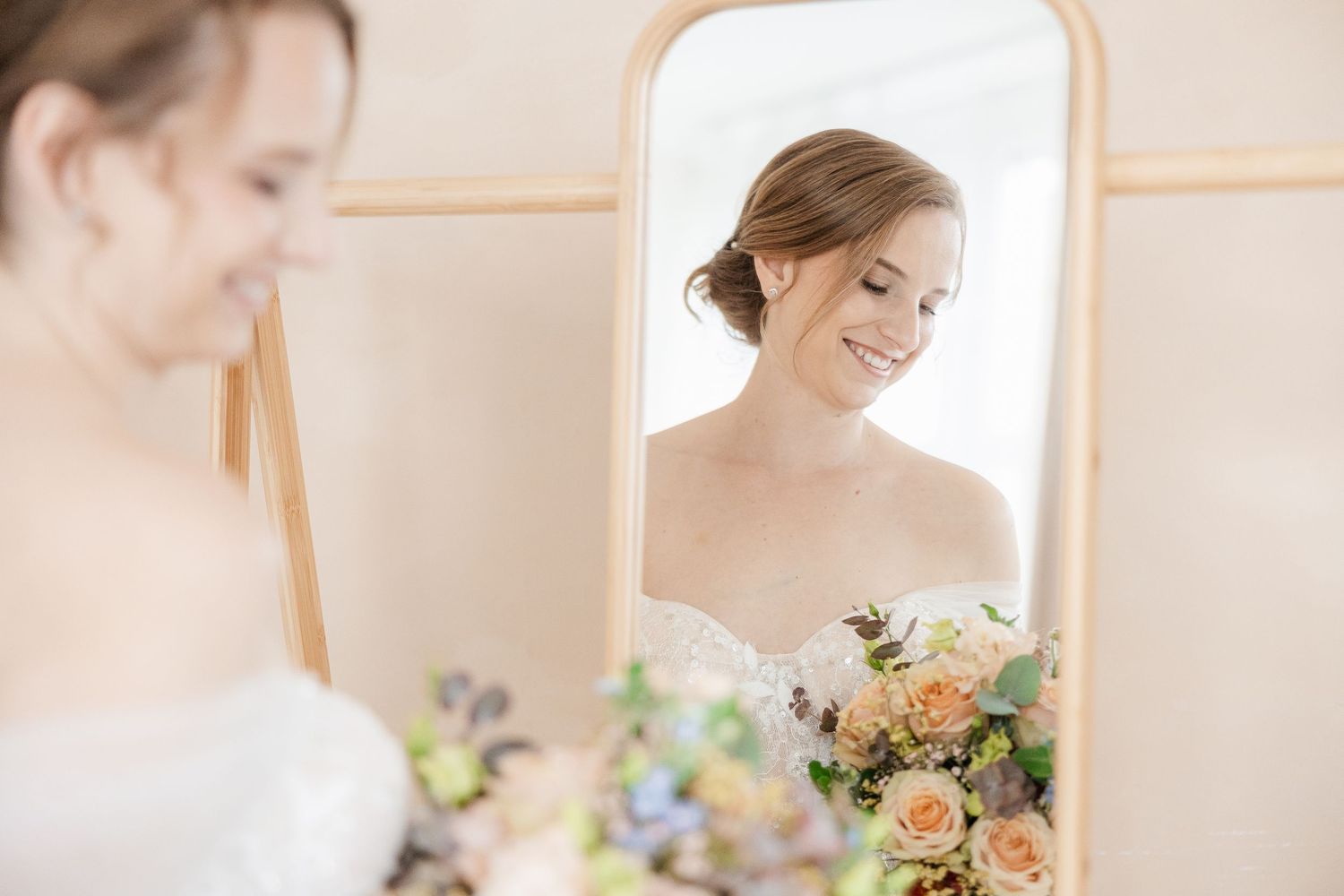 A bride in a white wedding gown holds a beautiful pastel bouquet of peach and pink flowers against a light background.