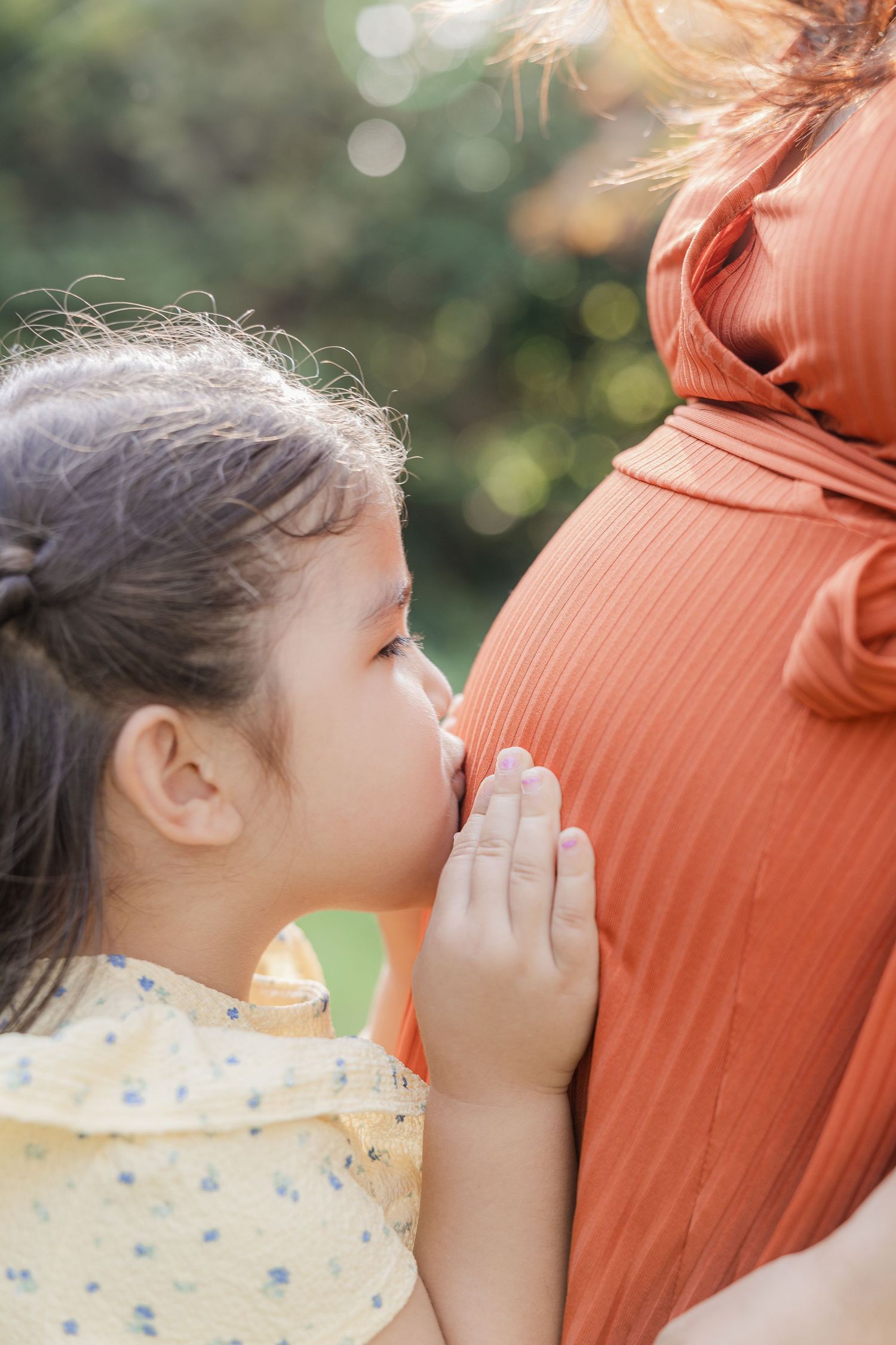 A series of photos showing a child in a yellow dress kissing a pregnant belly in an outdoor setting with soft bokeh lighting.
