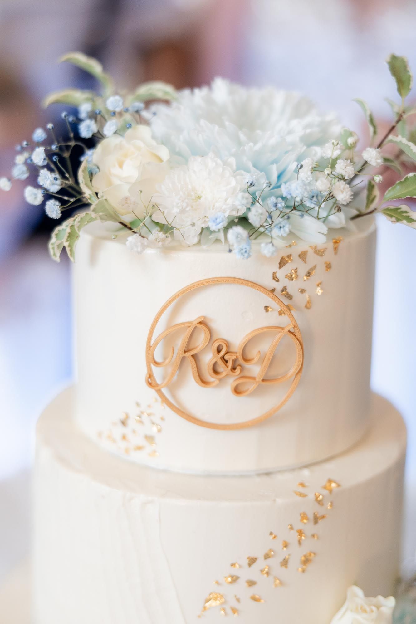 Elegant white tiered wedding cake with R&D monogram topper, gold accents, and soft blue and white floral decorations.