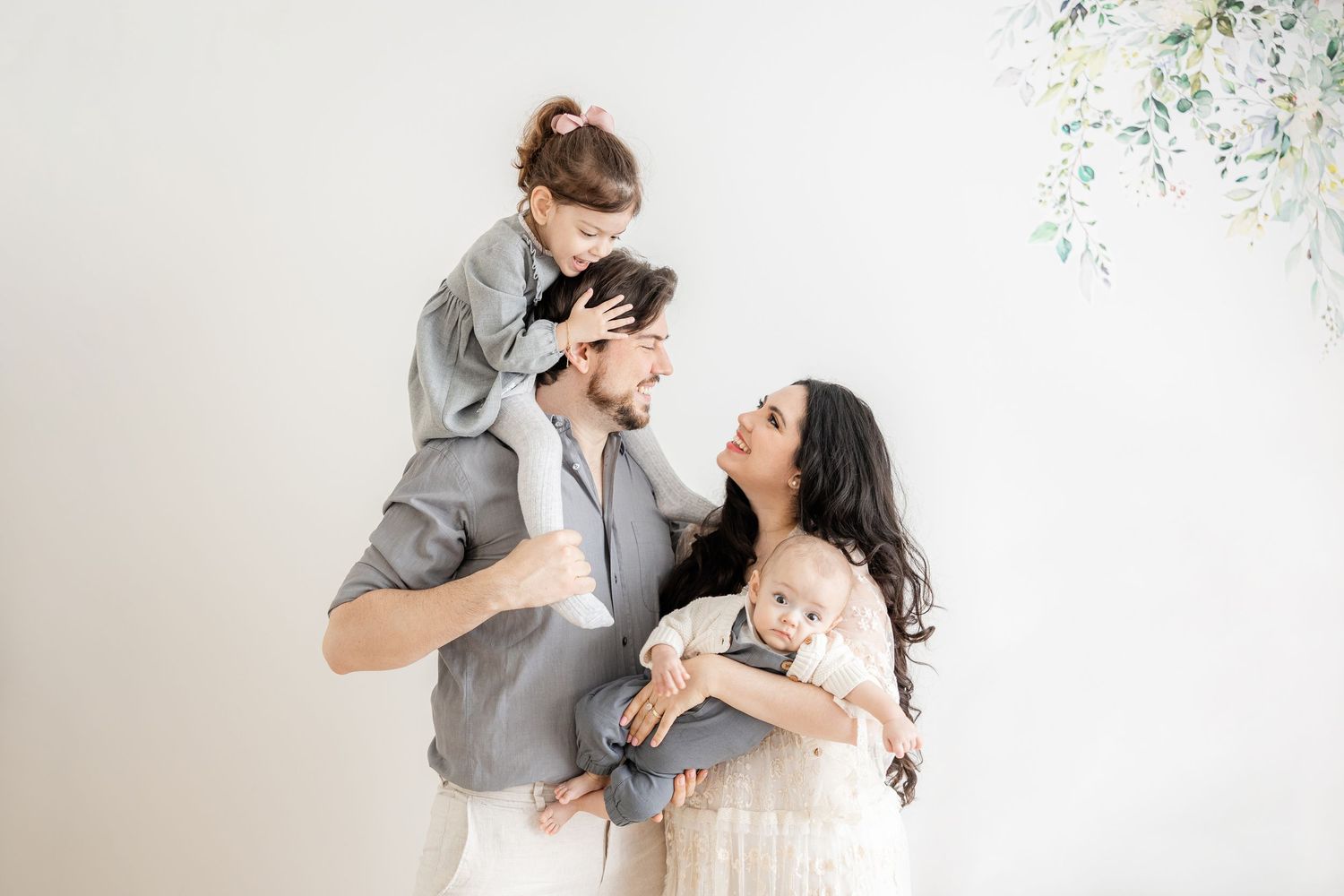 A joyful family in light neutral colors poses together against a white wall with delicate floral decor. Gränichen, Aargau, Schweiz