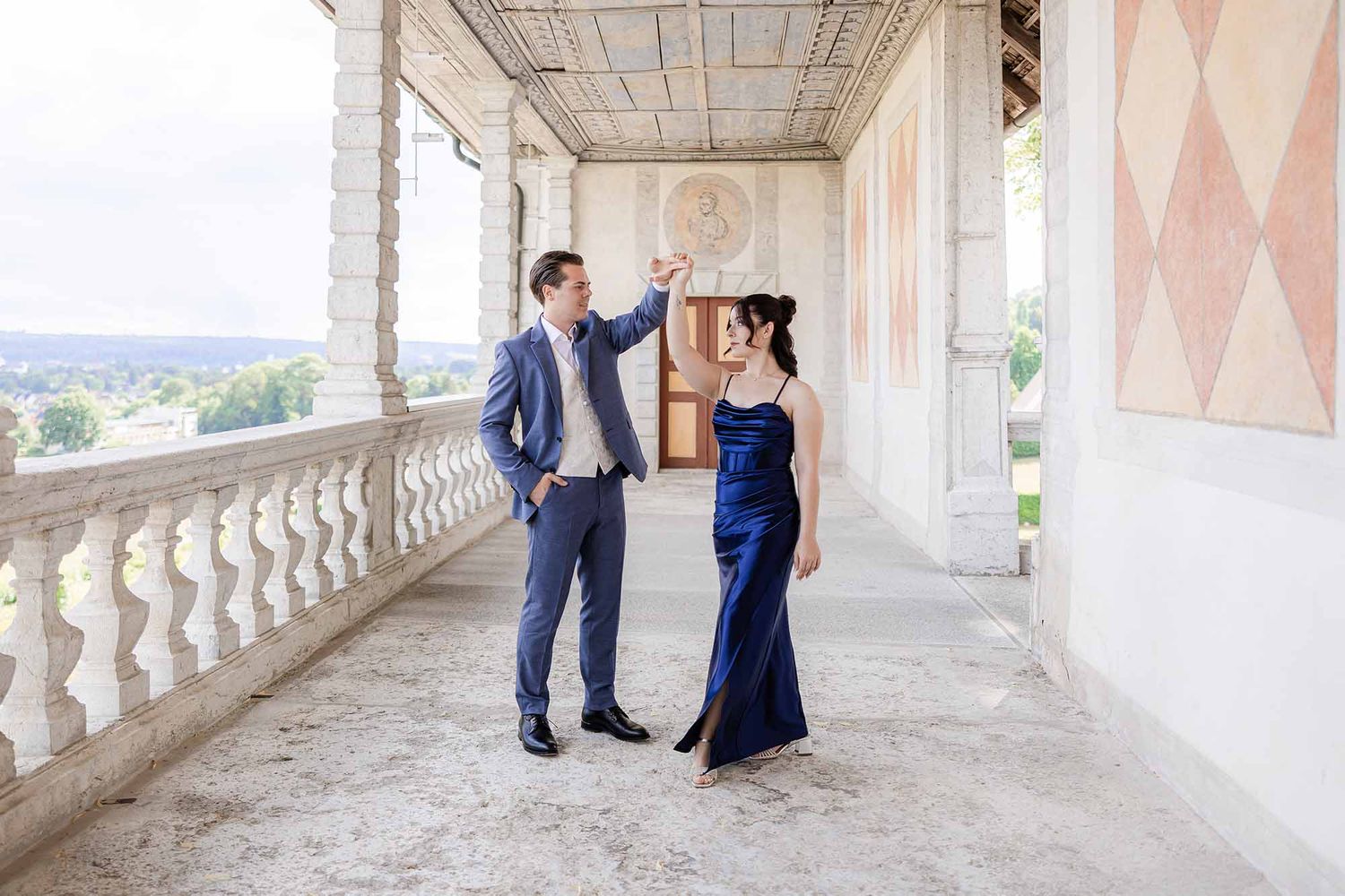 Elegant couple in formal attire dancing on a grand balcony with scenic views and ornate architecture.