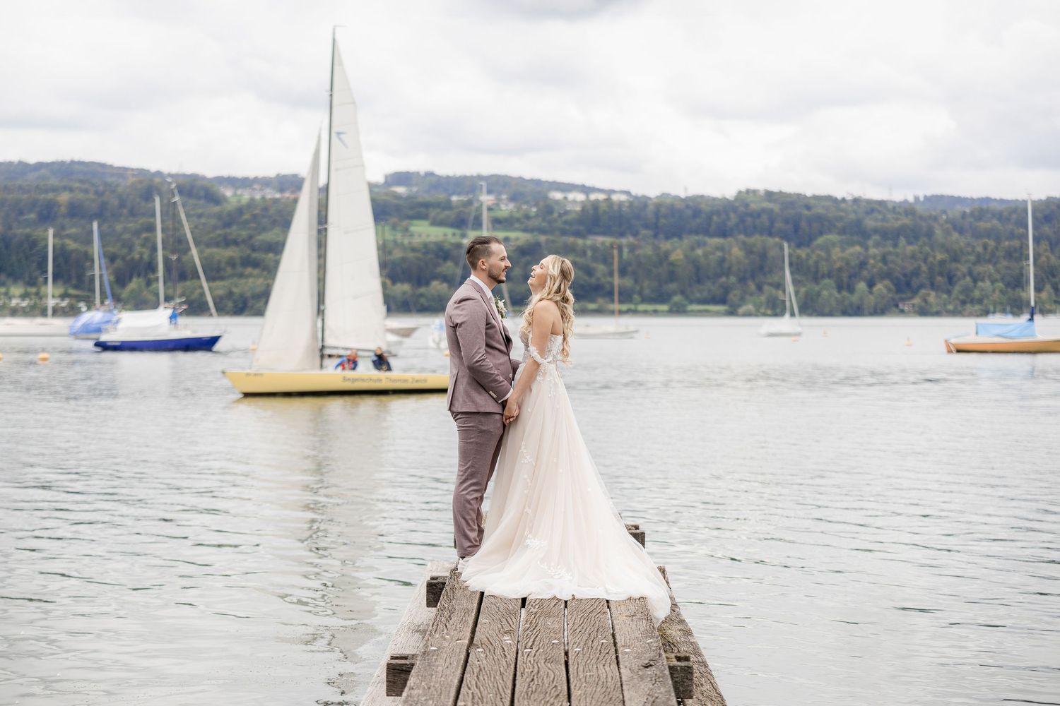 A couple stands at the end of a wooden dock overlooking a lake with sailboats in the distance on an overcast day.