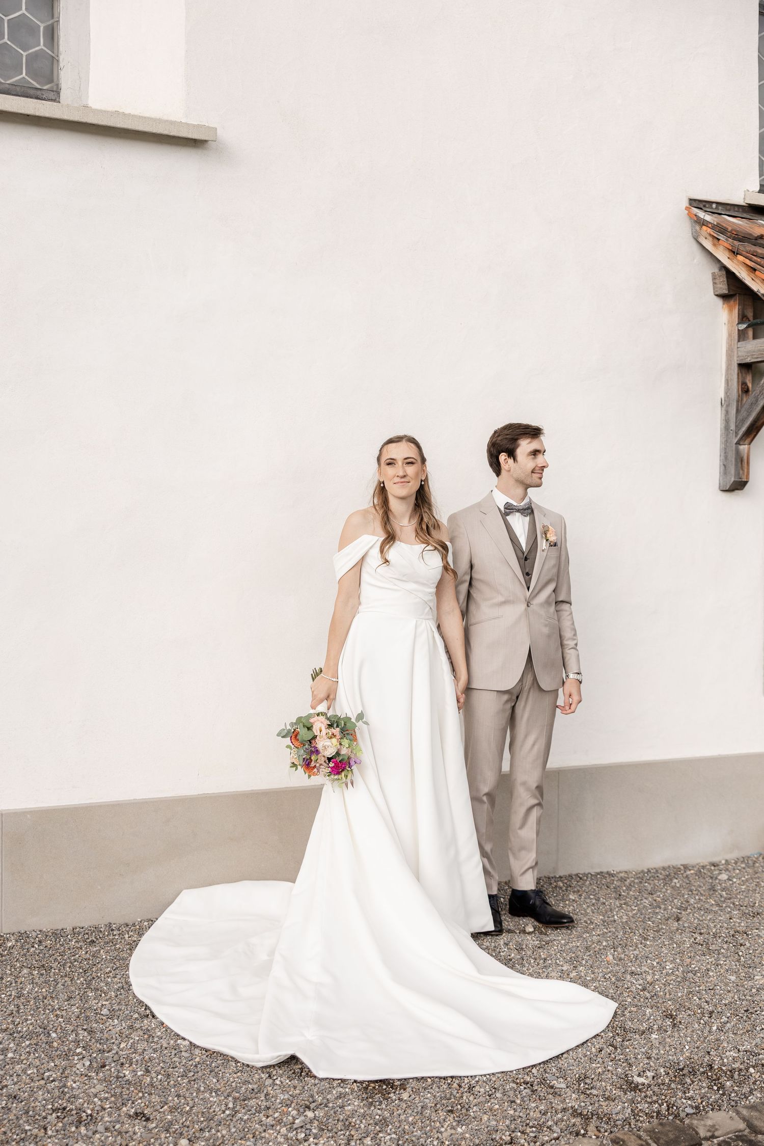 A bride and groom in a white dress and gray suit pose together against a white wall in a romantic wedding photo series.