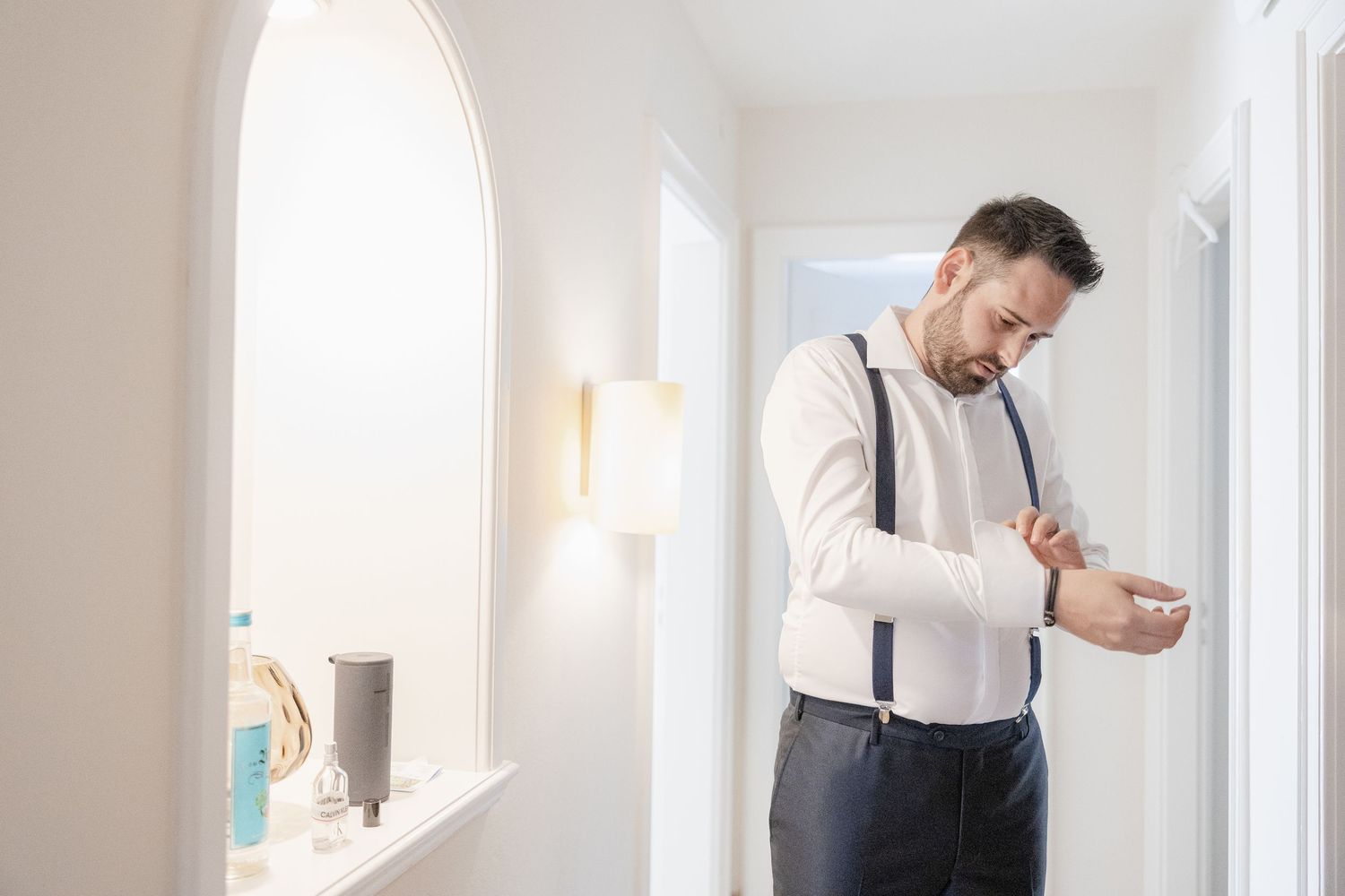 A person in a white shirt and suspenders adjusts their outfit while standing in a bright interior space.