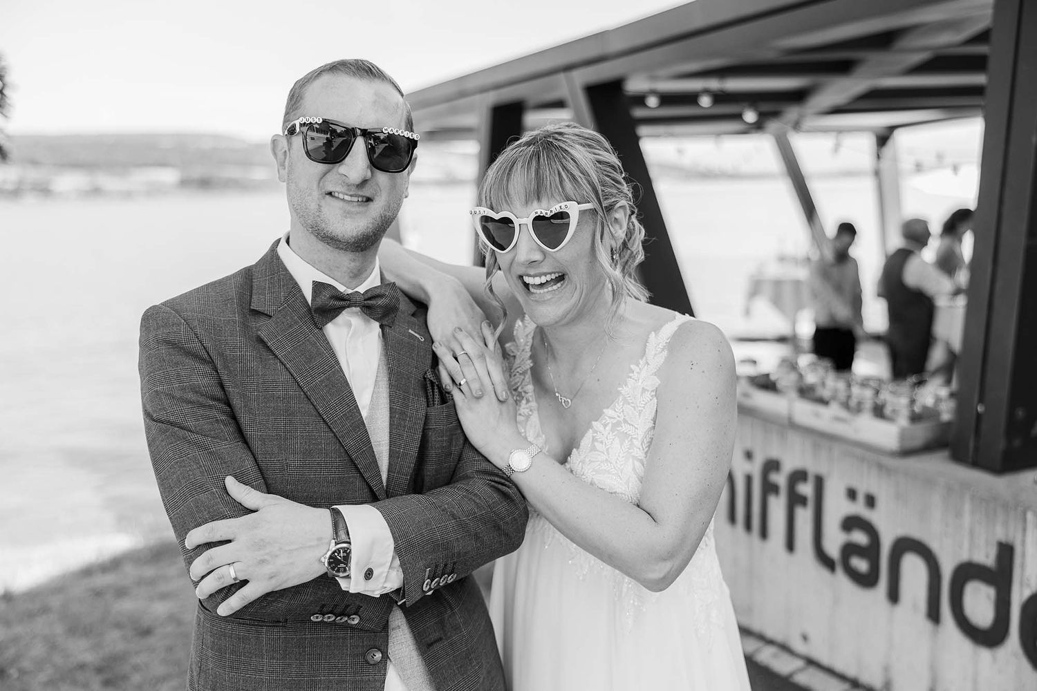Black and white photo of a smiling bride in heart sunglasses and groom in bow tie posing together at an outdoor wedding.