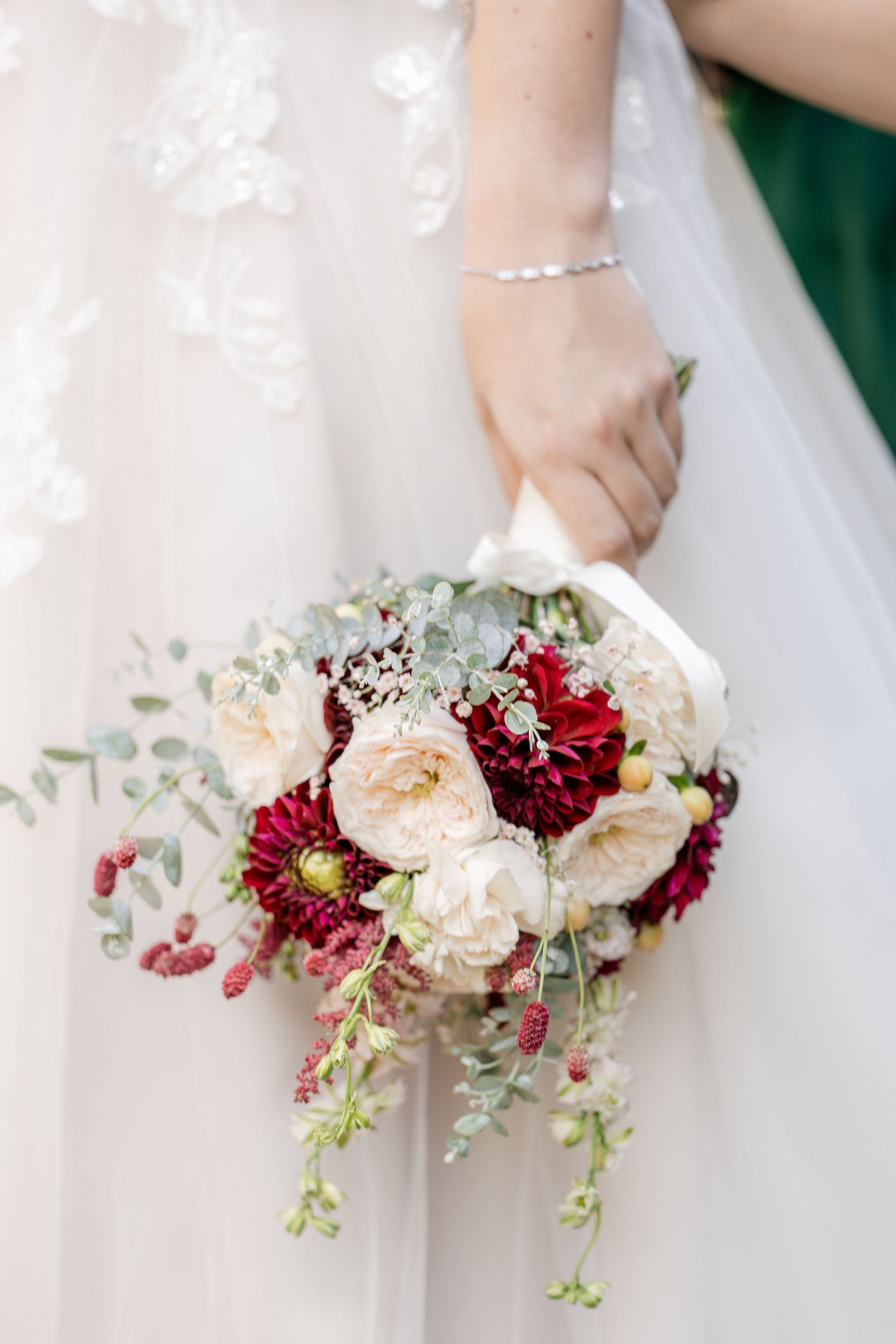 A bridal bouquet featuring cream garden roses, deep red dahlias, and cascading eucalyptus against a white wedding dress.