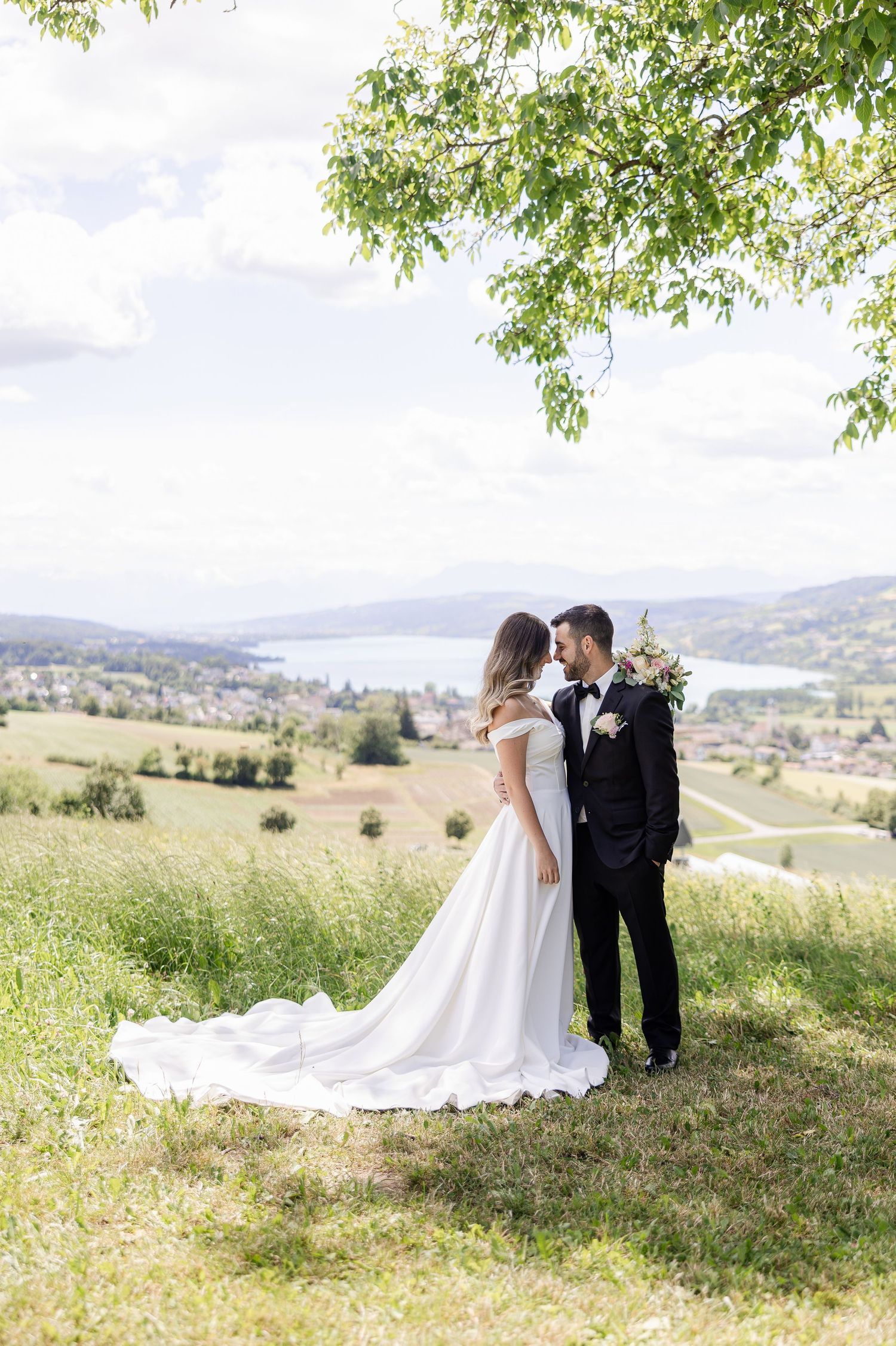 Bride and groom share a romantic moment on a green hillside with a scenic lake and valley view on their wedding day.