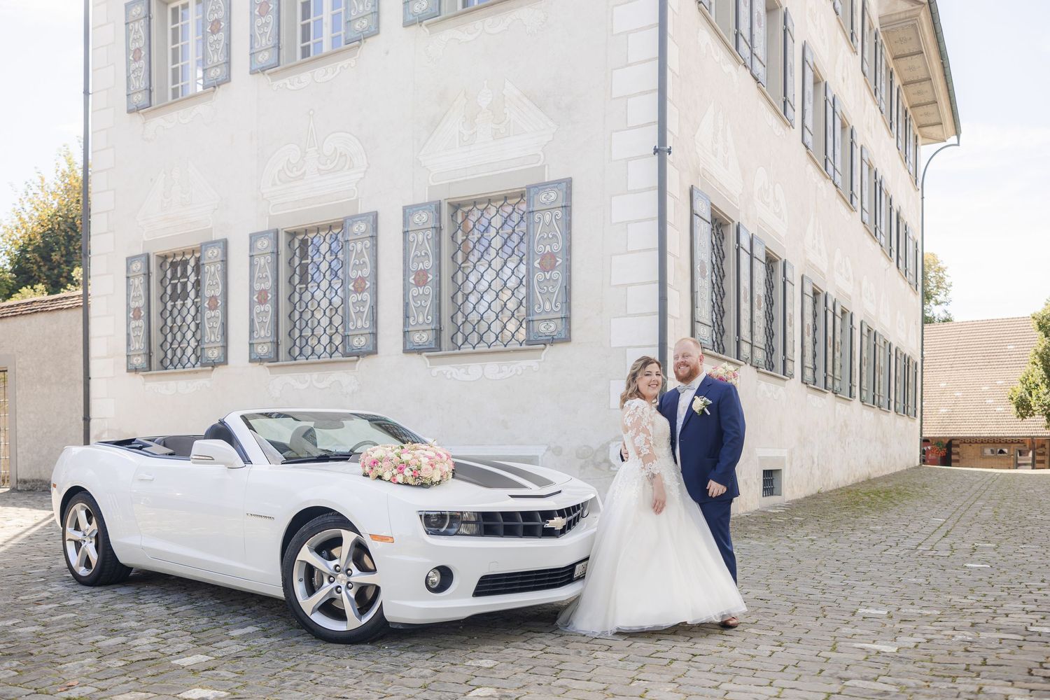 White Chevrolet Camaro convertible parked in front of an elegant white building with blue shutters.