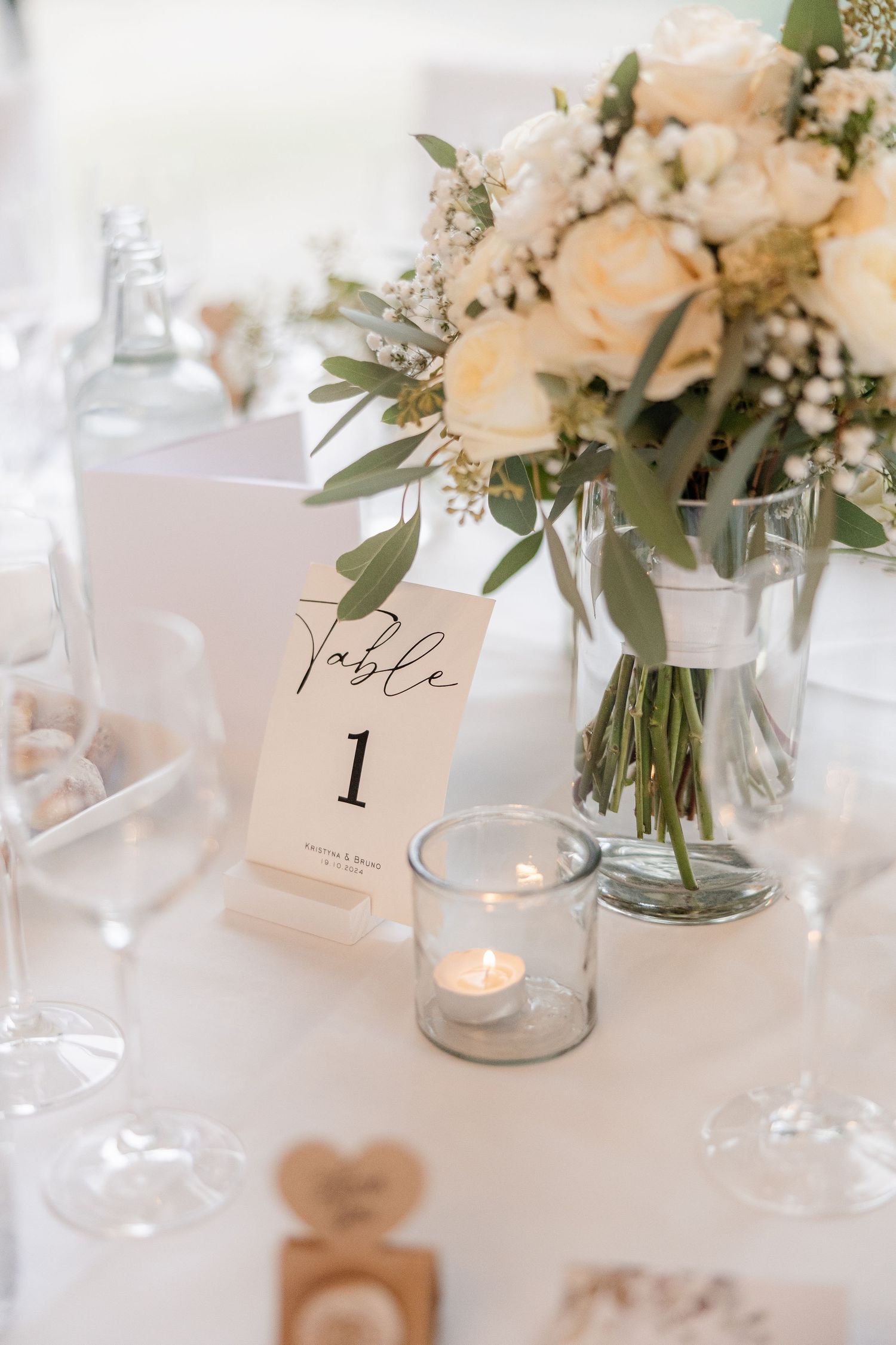 Elegant wedding table decor with white floral arrangements in glass vases, tea lights, and table number cards on crisp linens.