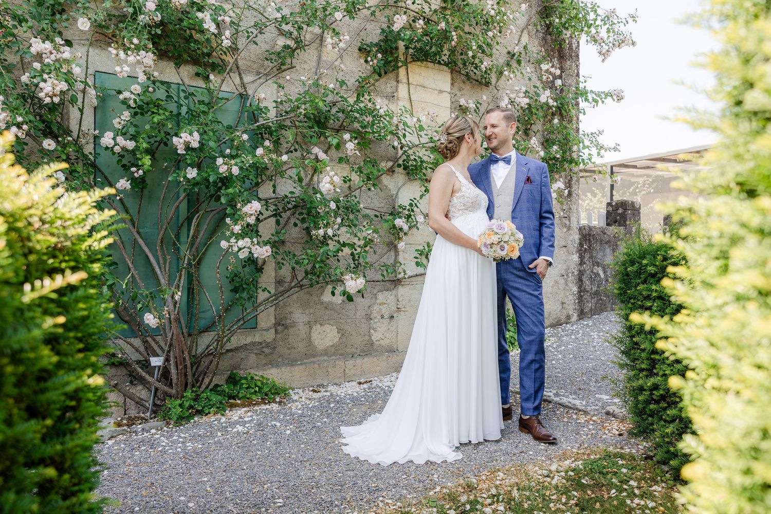 Bride in a flowing white gown and groom in a blue suit share a tender moment on a gravel path beside a stone wall with climbing white roses.