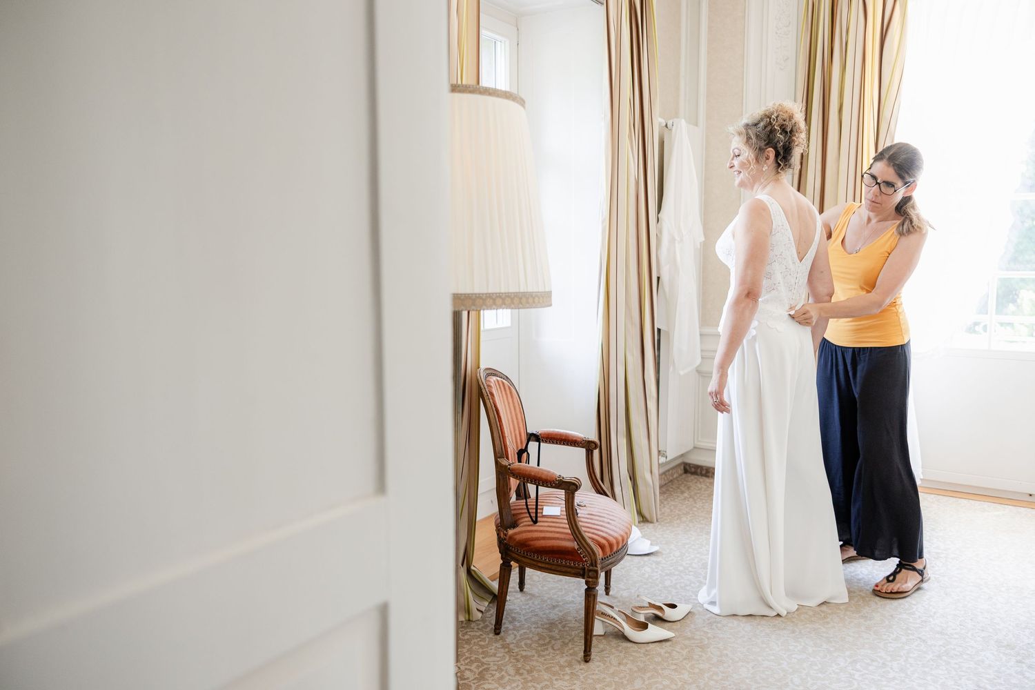Bridesmaid in yellow dress helping bride with her white wedding gown in an elegant hotel room.