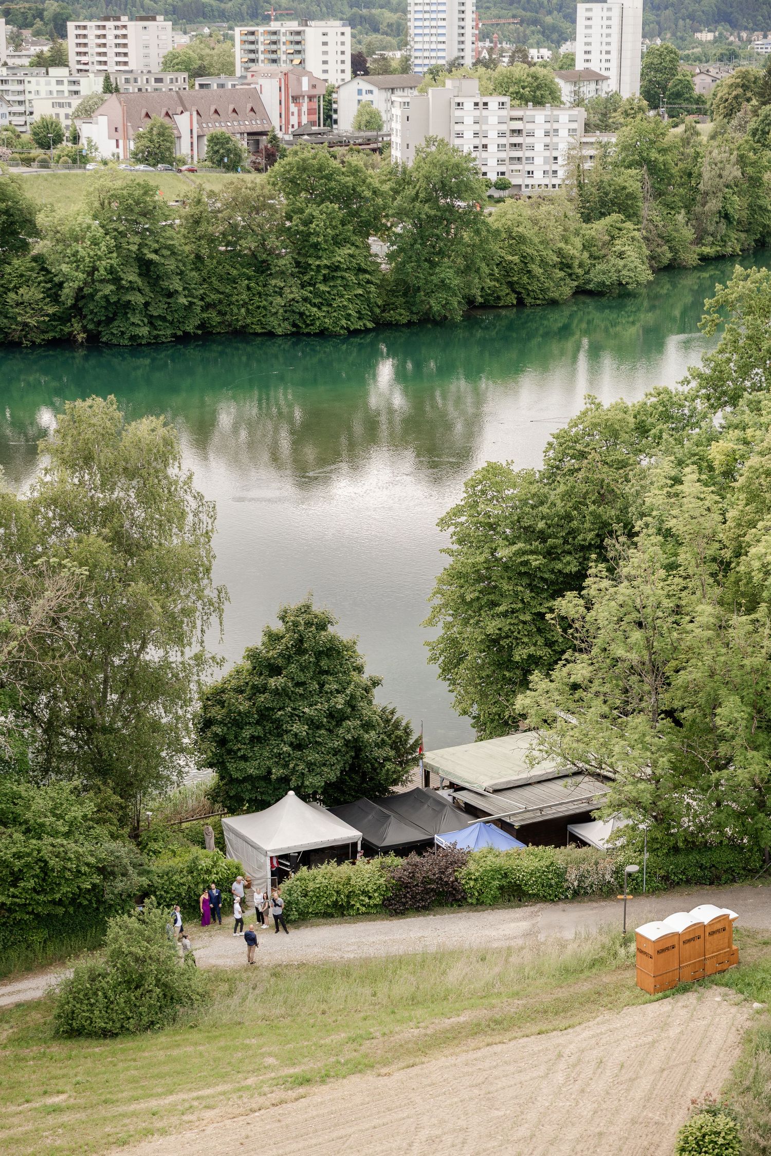 Scenic riverside park with green trees, walking paths and apartment buildings visible in the distance. Fischerhütte Wettingen, Baden, Aargau