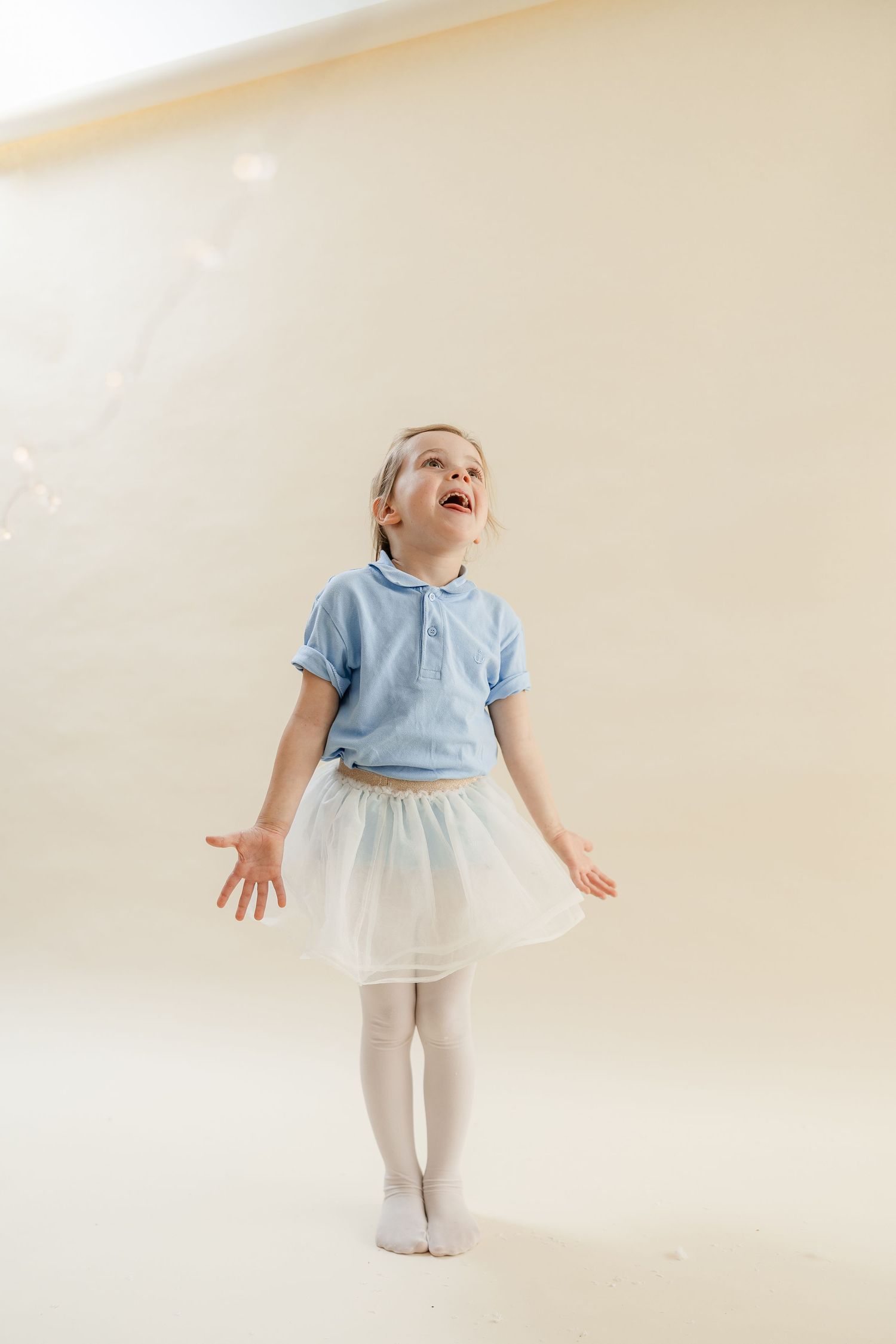 Young ballet dancer in light blue leotard and white tutu performs graceful poses in a bright warm studio setting.