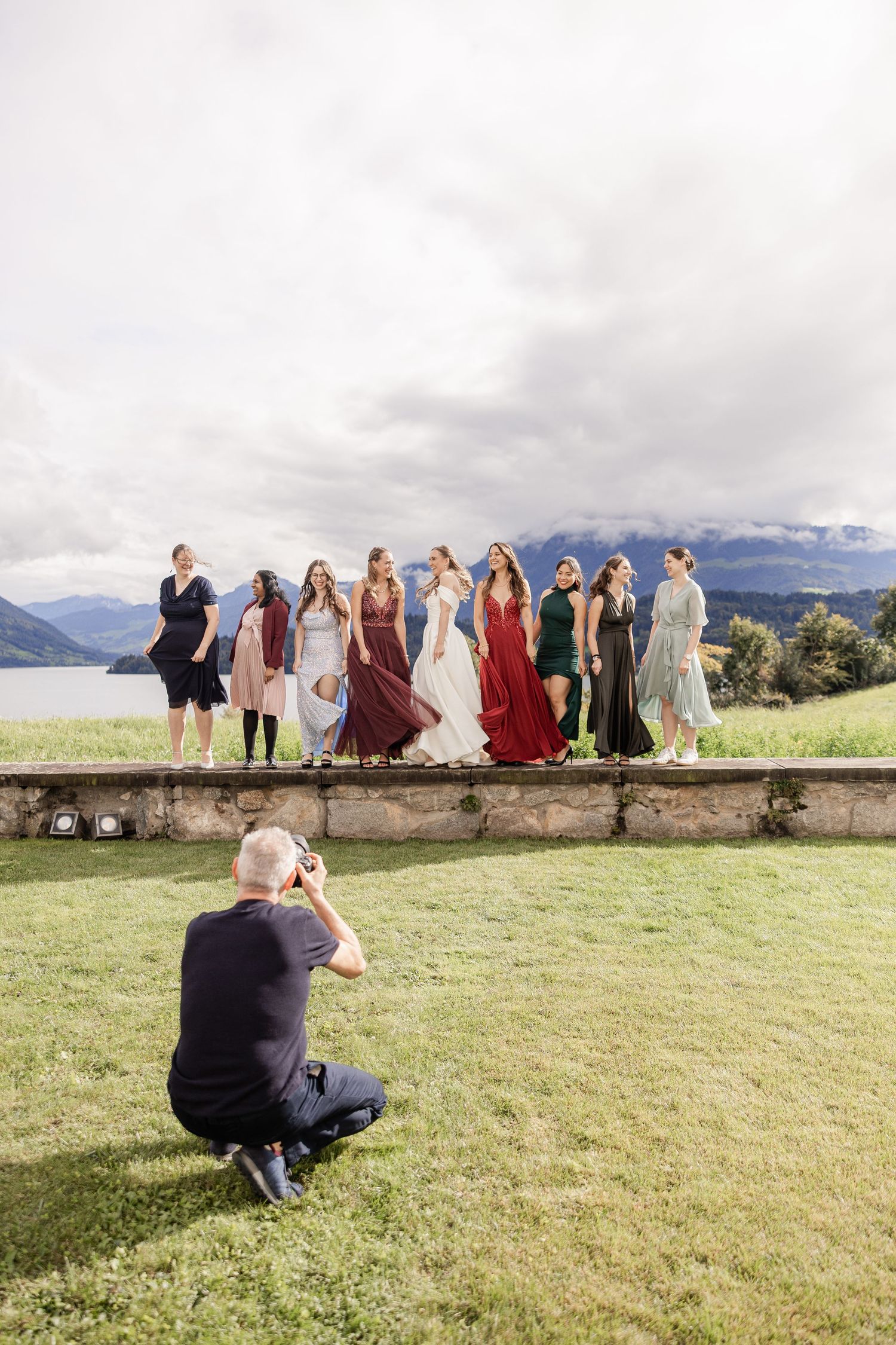 Behind the scenes photographer captures a wedding party sitting on stone wall overlooking mountain landscape.