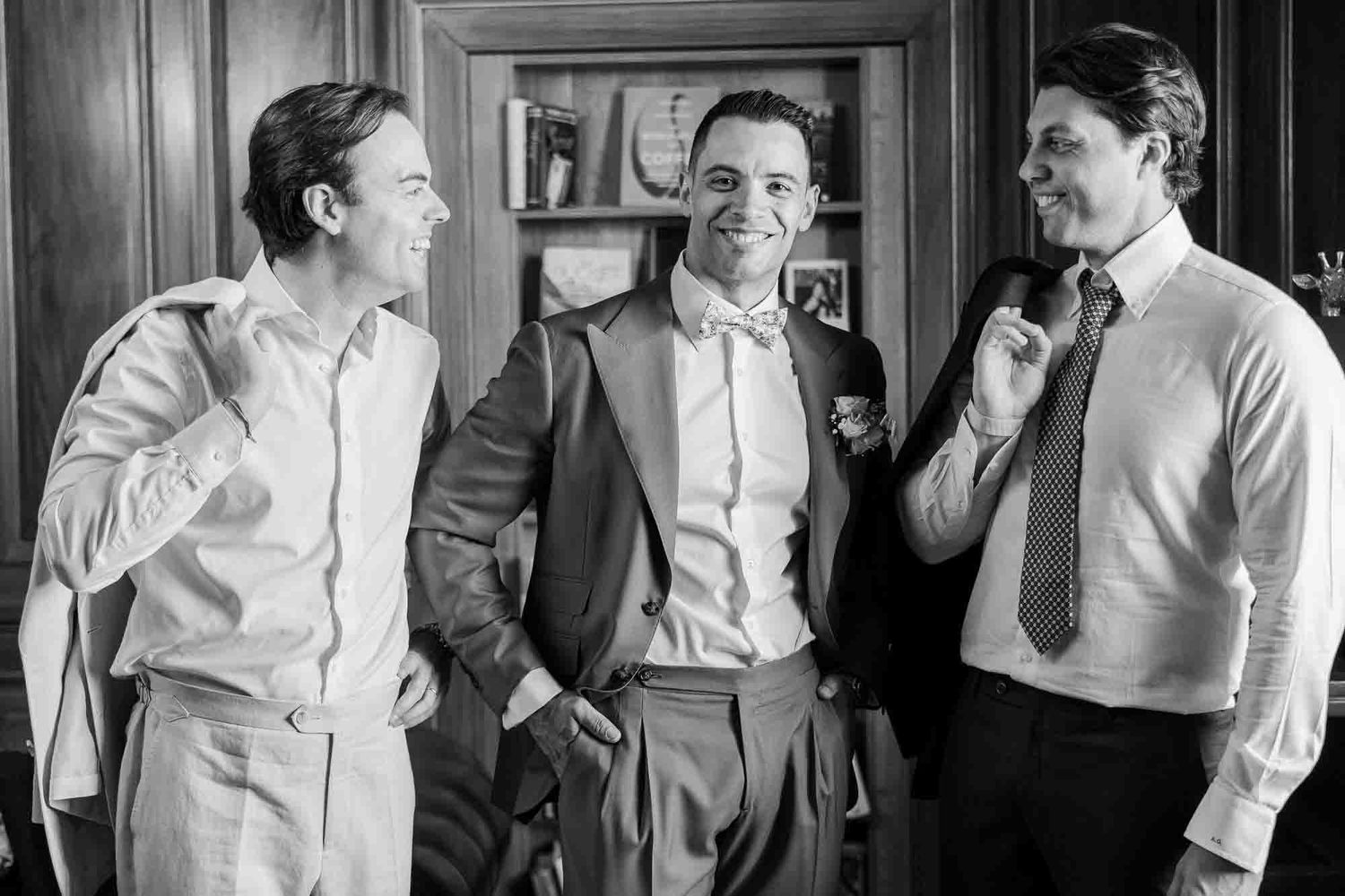Three men in formal attire laugh together, with the groom wearing a bow tie flanked by his groomsmen.