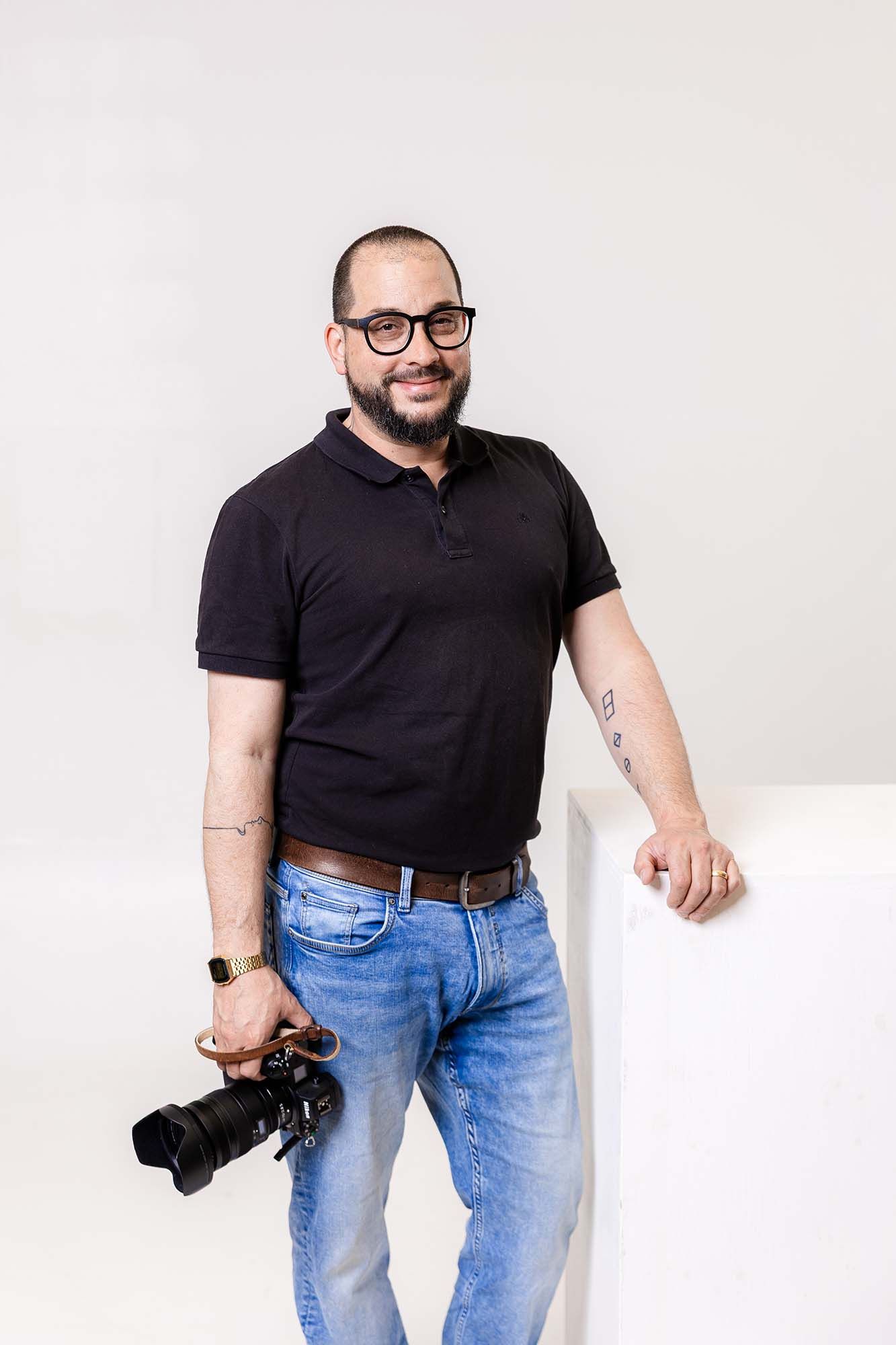 A photographer in a black t-shirt and jeans poses with a camera against a white background.