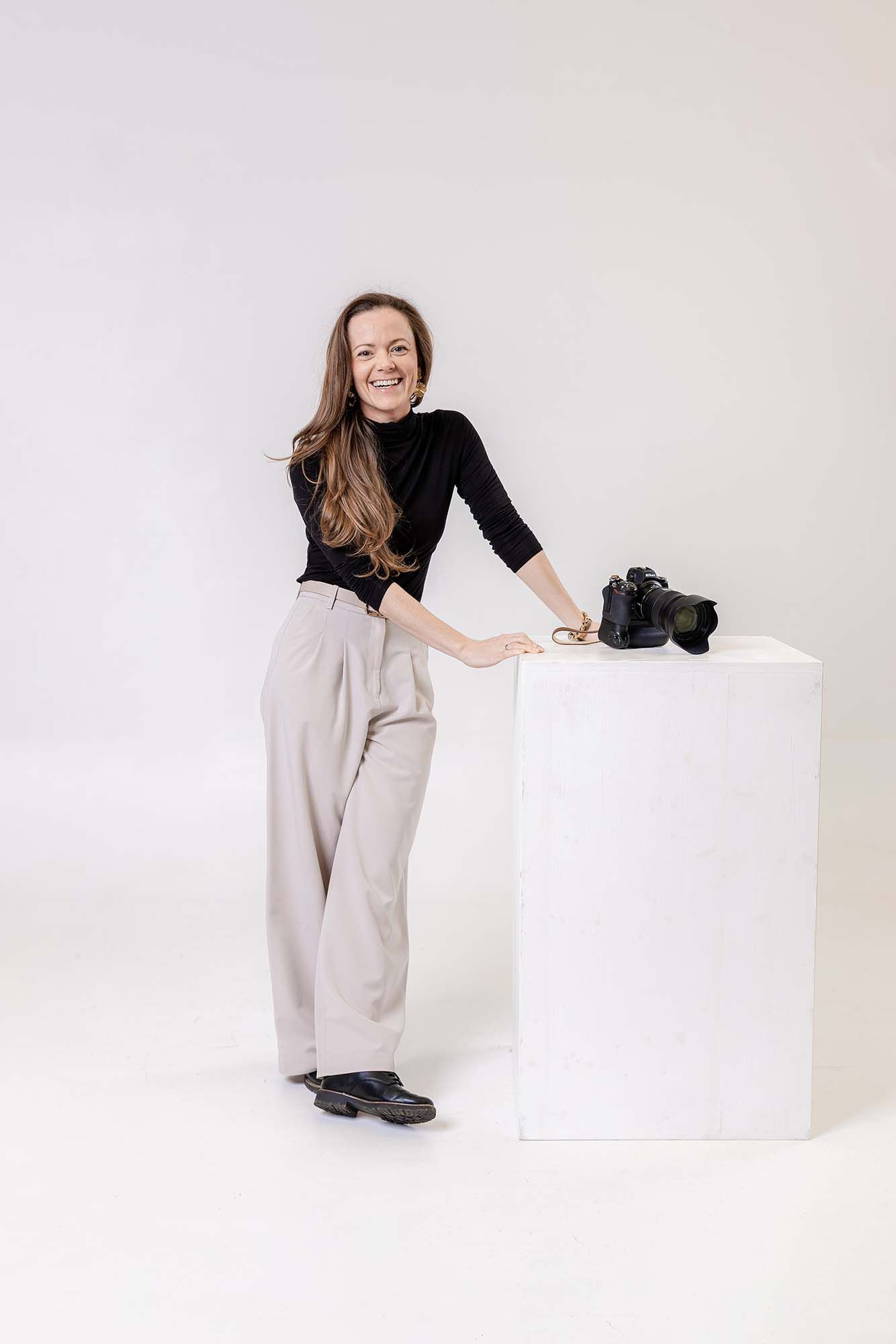 Professional photographer in black and beige outfit poses with camera equipment against white studio backdrop.