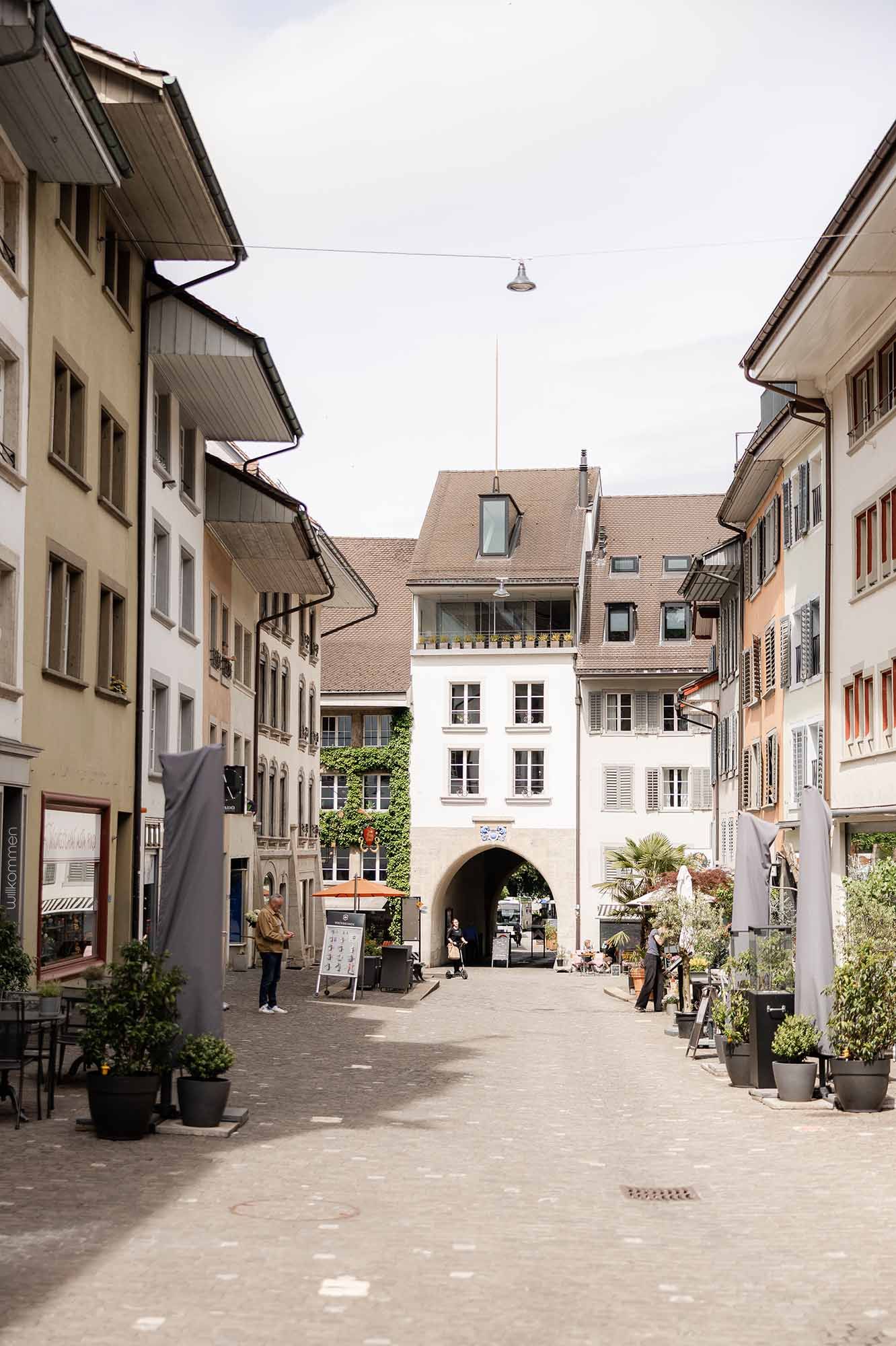 Charming European pedestrian street with historic gate building, cobblestone path, and outdoor cafes on a sunny day.