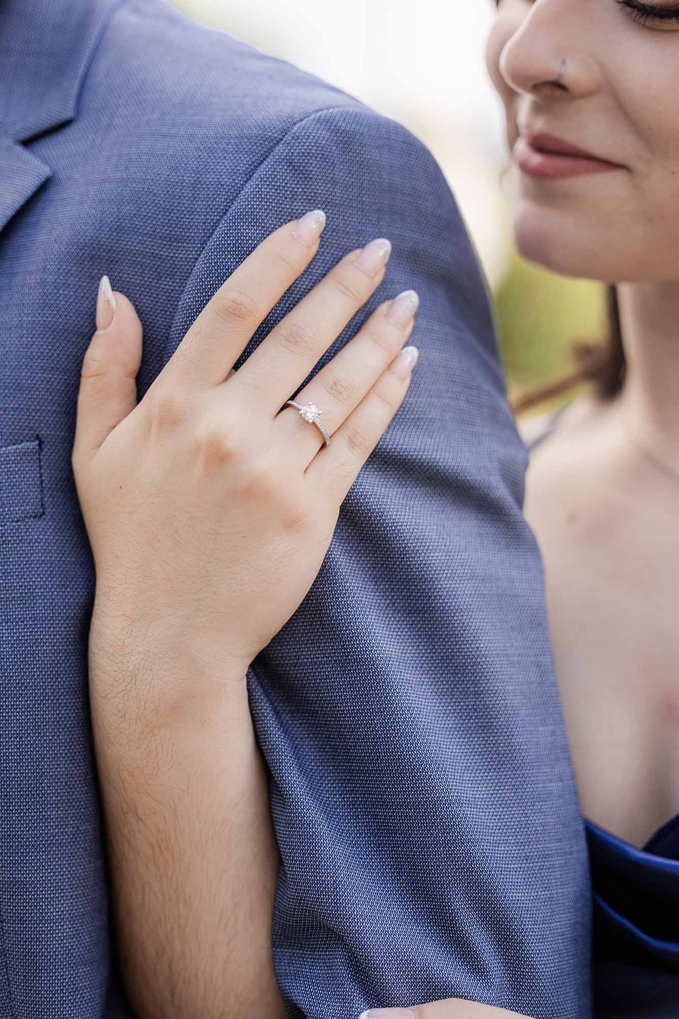 Woman wearing a diamond engagement ring rests her hand on a man's blue suit jacket shoulder.