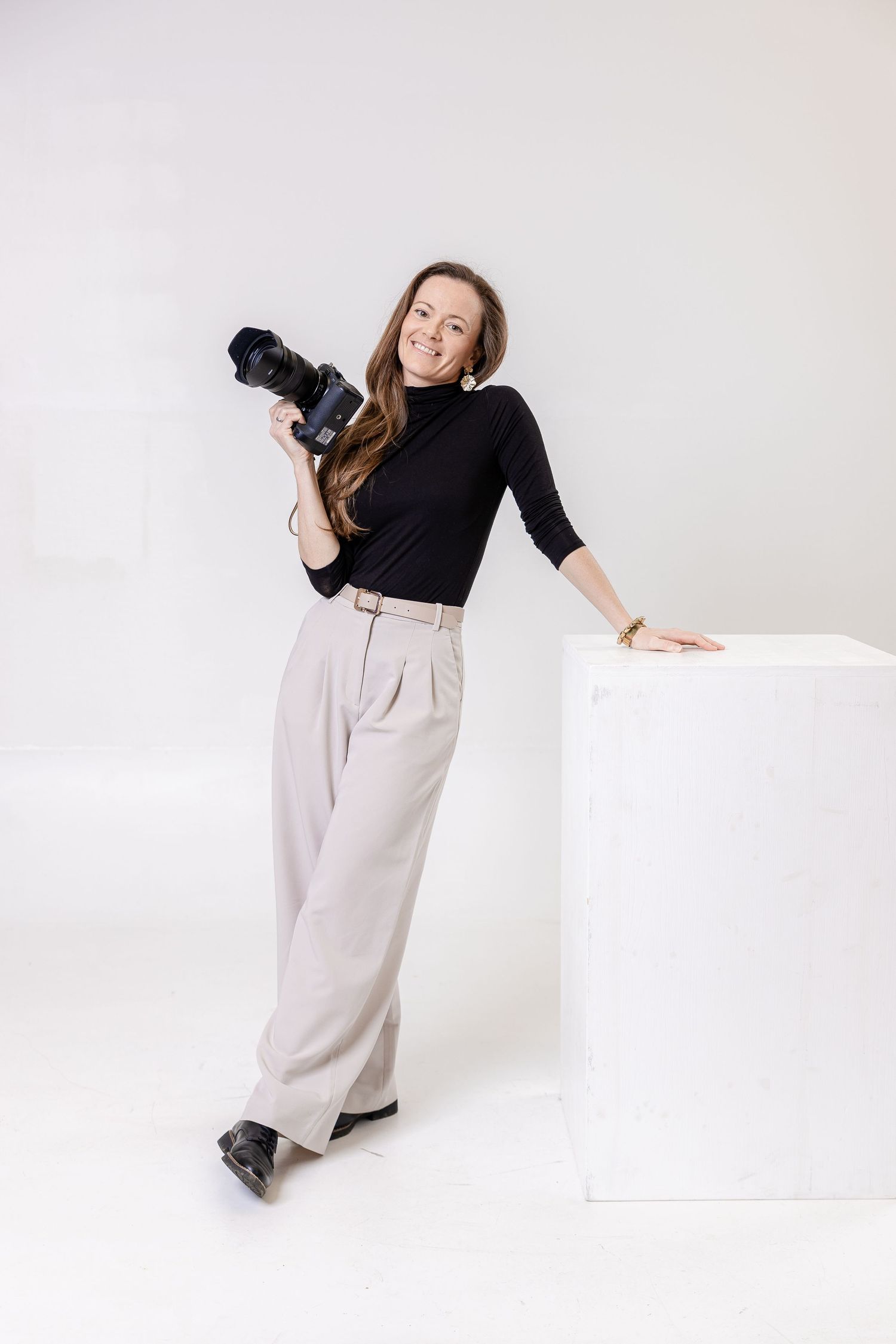 Smiling female photographer in black turtleneck and wide-leg pants holding a camera, leaning on white cube in studio.