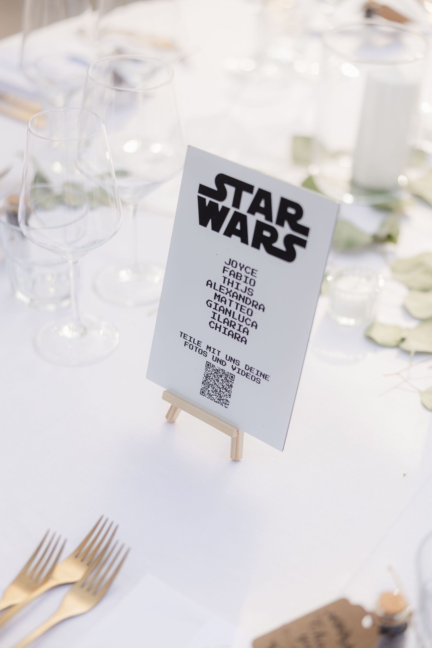 A Star Wars themed wedding table sign displayed on a small easel among wine glasses and place settings.