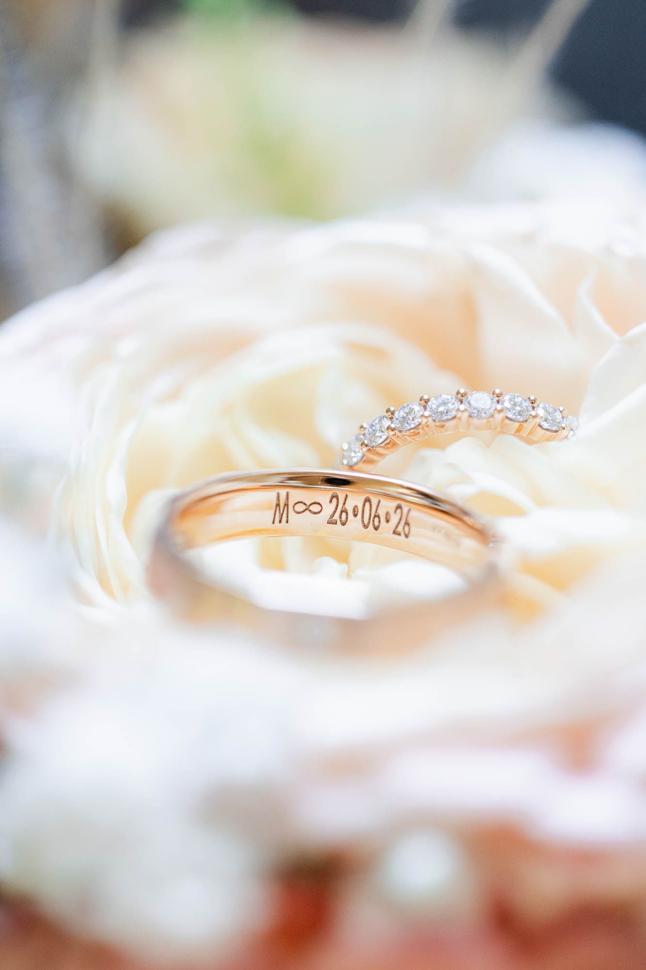 Gold wedding bands and diamond ring resting on a white rose, engraved with M∞26•06•24.