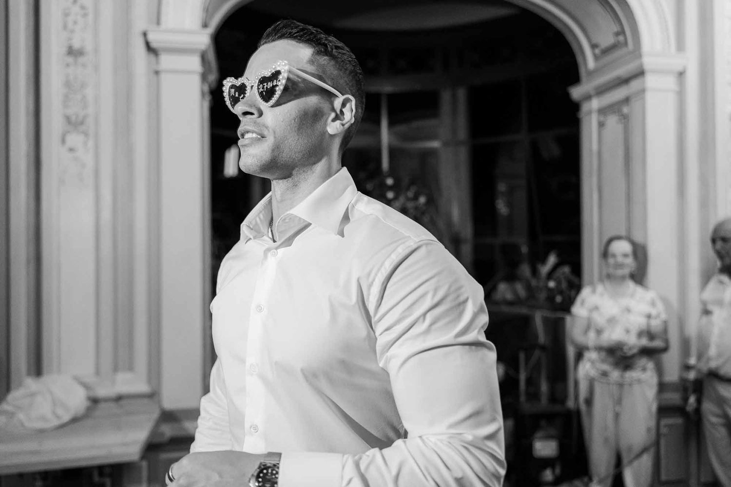 Black and white photo of a stylish man wearing heart-shaped sunglasses and a white shirt at an elegant venue.