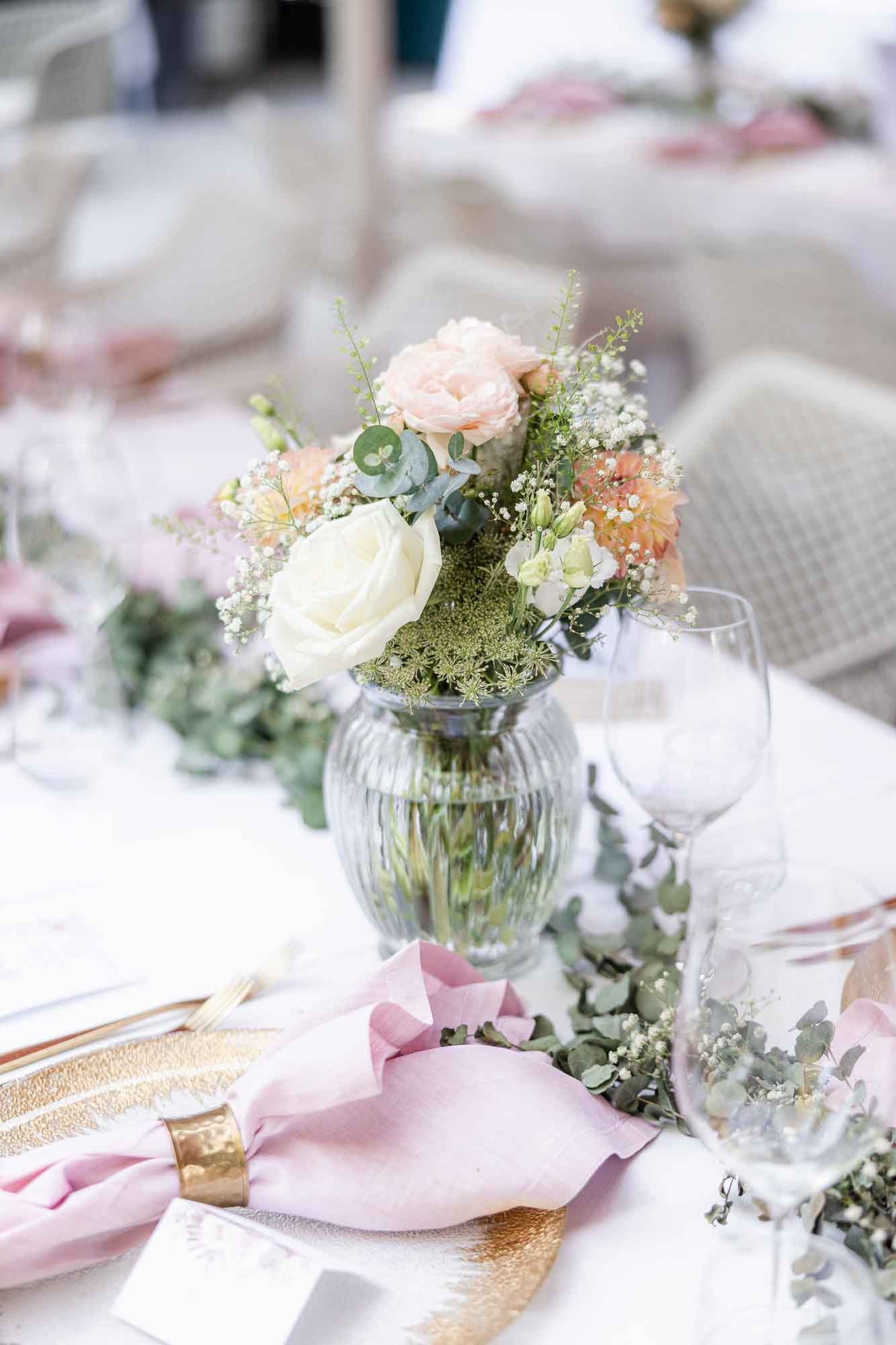 Elegant wedding table setting with pink roses, white flowers, blush napkins, and gold chargers in a glass vase centerpiece.