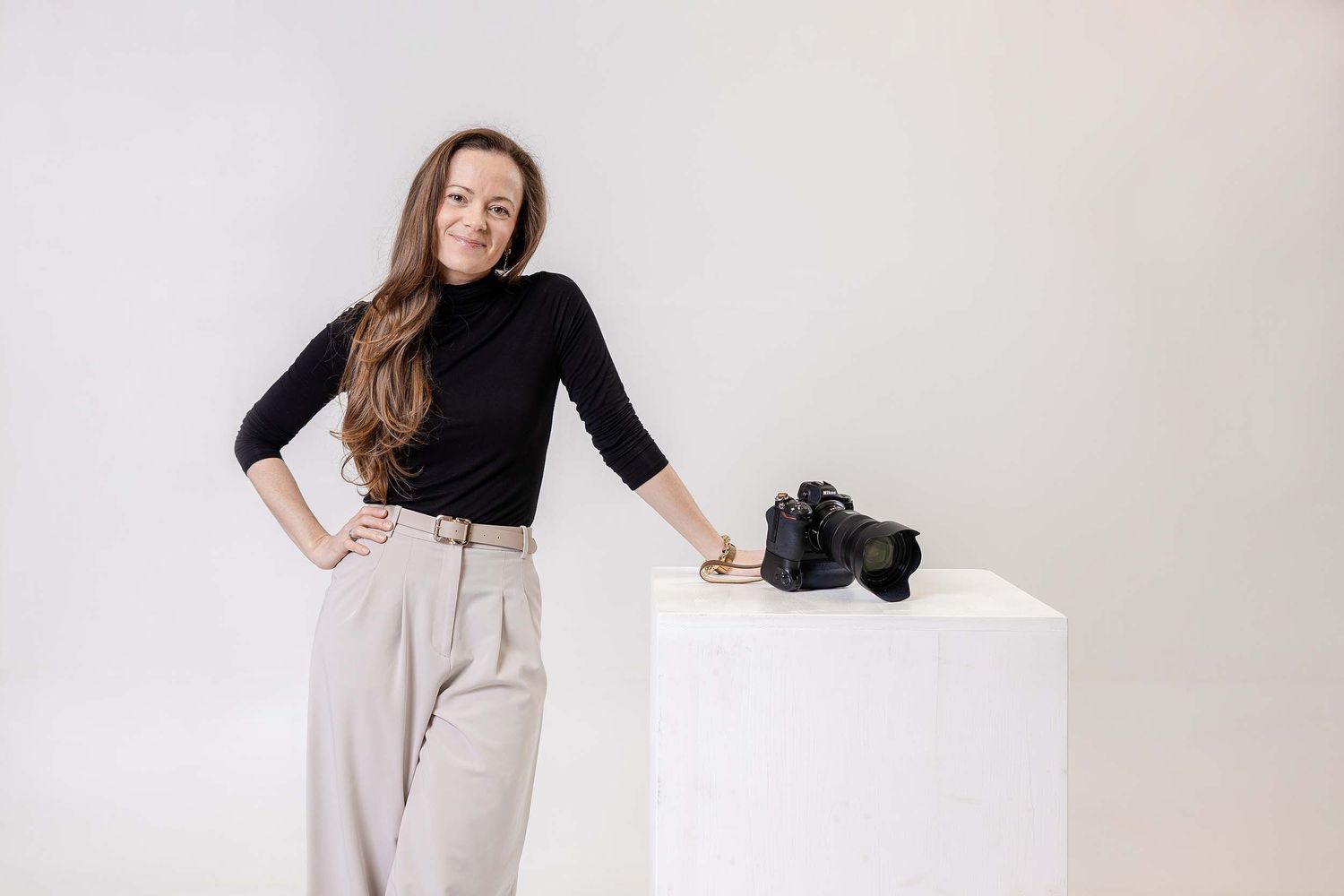 Professional fashion model wearing black turtleneck and beige pants poses in studio against white background.