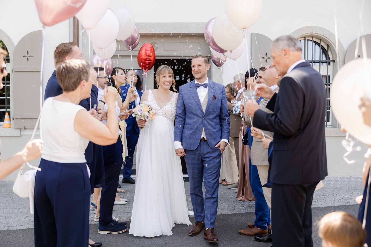 Newlywed couple walks through cheering guests holding balloons outside a church after their wedding ceremony.