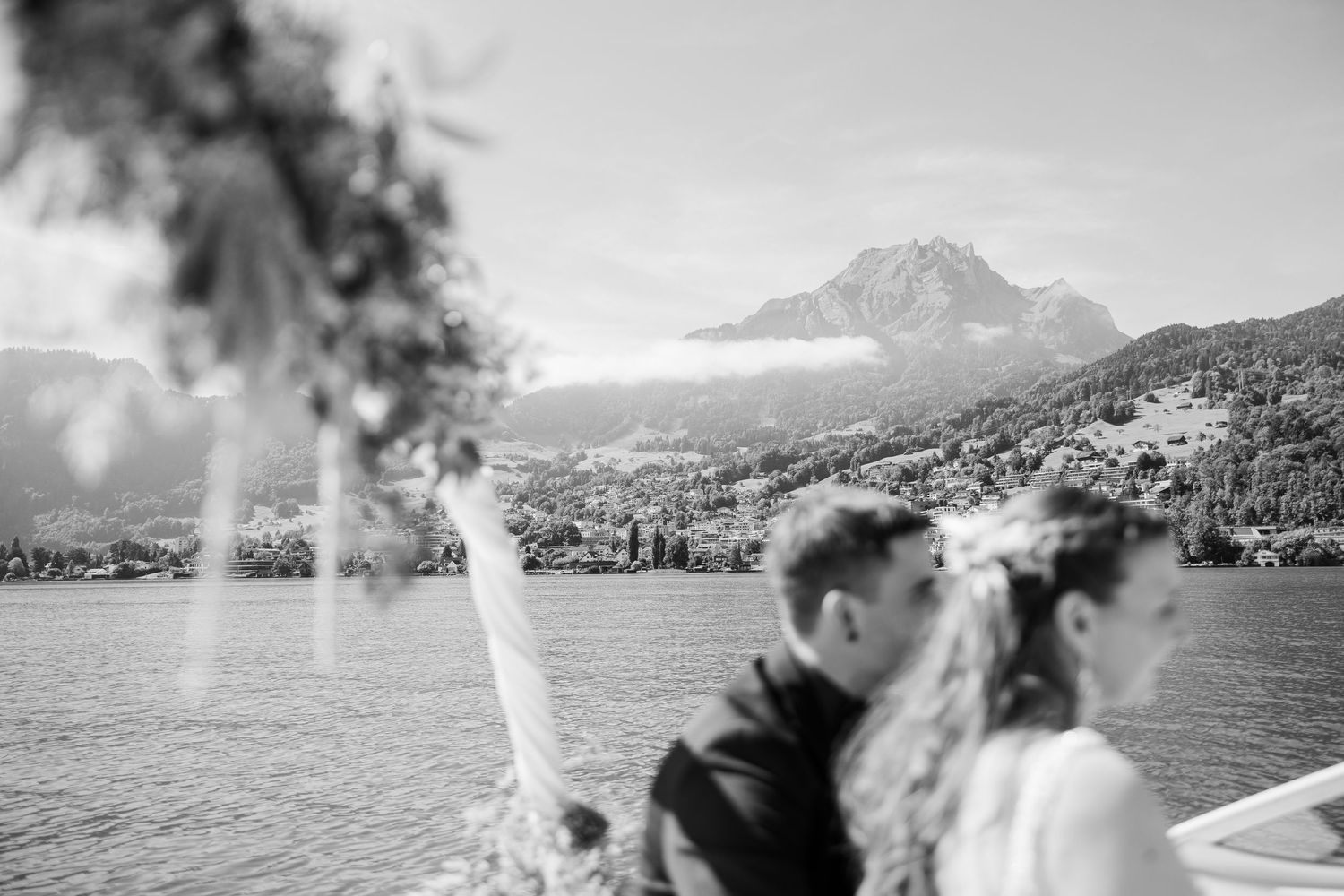 Black and white photo of a bride and groom on a boat with a stunning mountain lake backdrop.