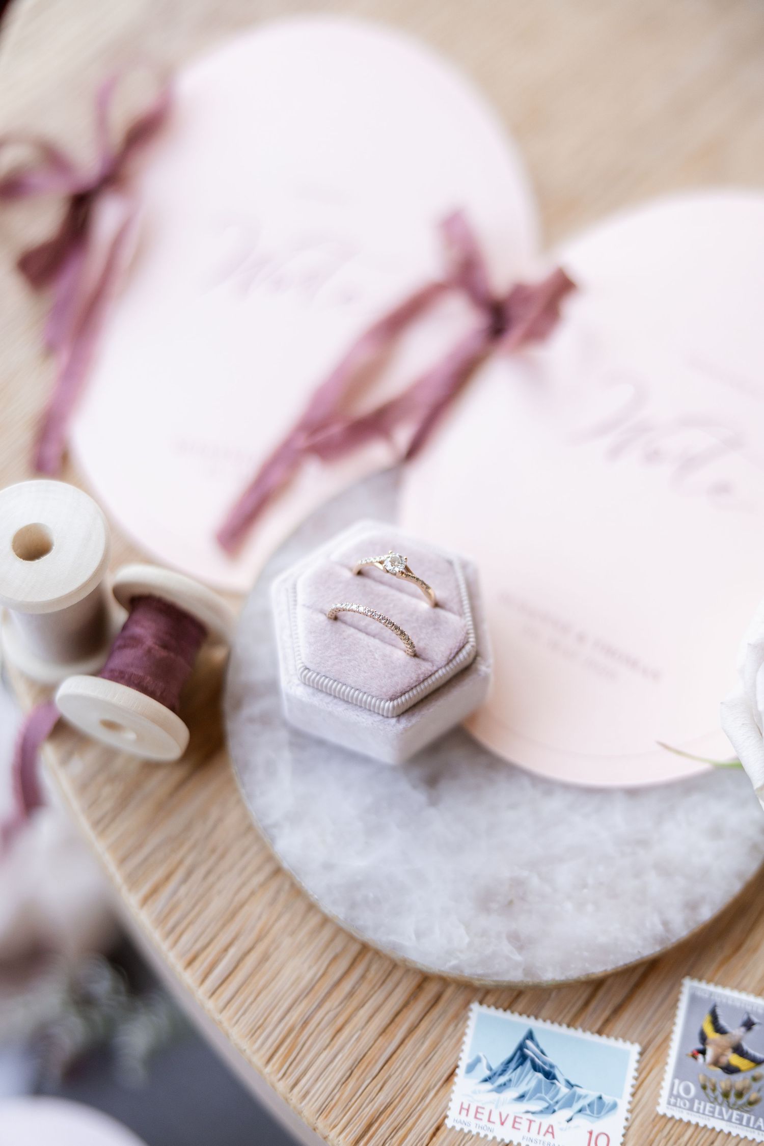 Elegant wedding flat lay with gold rings in velvet box, pink stationery, ribbon spools, and Swiss stamps on wooden surface.