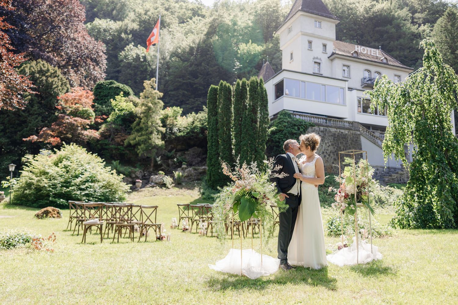 Bride and groom embrace during outdoor wedding ceremony on lush green lawn with floral arch and elegant villa backdrop.