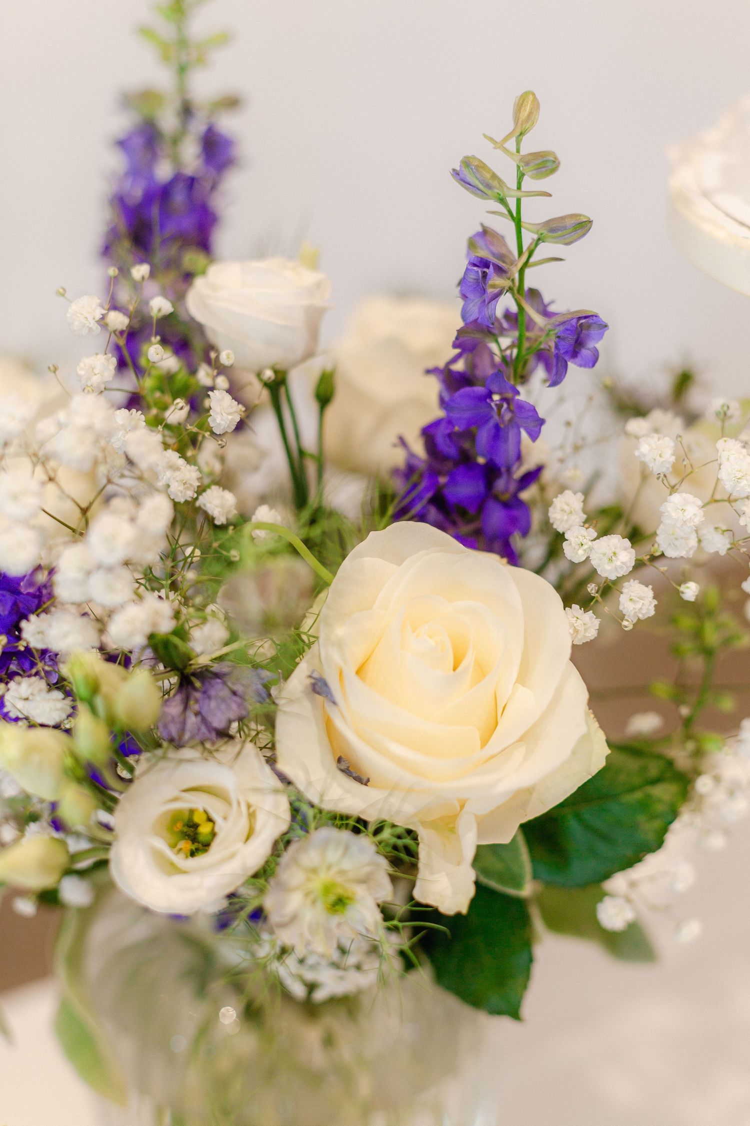 Close-up view of elegant cream roses and purple delphinium flowers with baby's breath in a wedding floral arrangement.