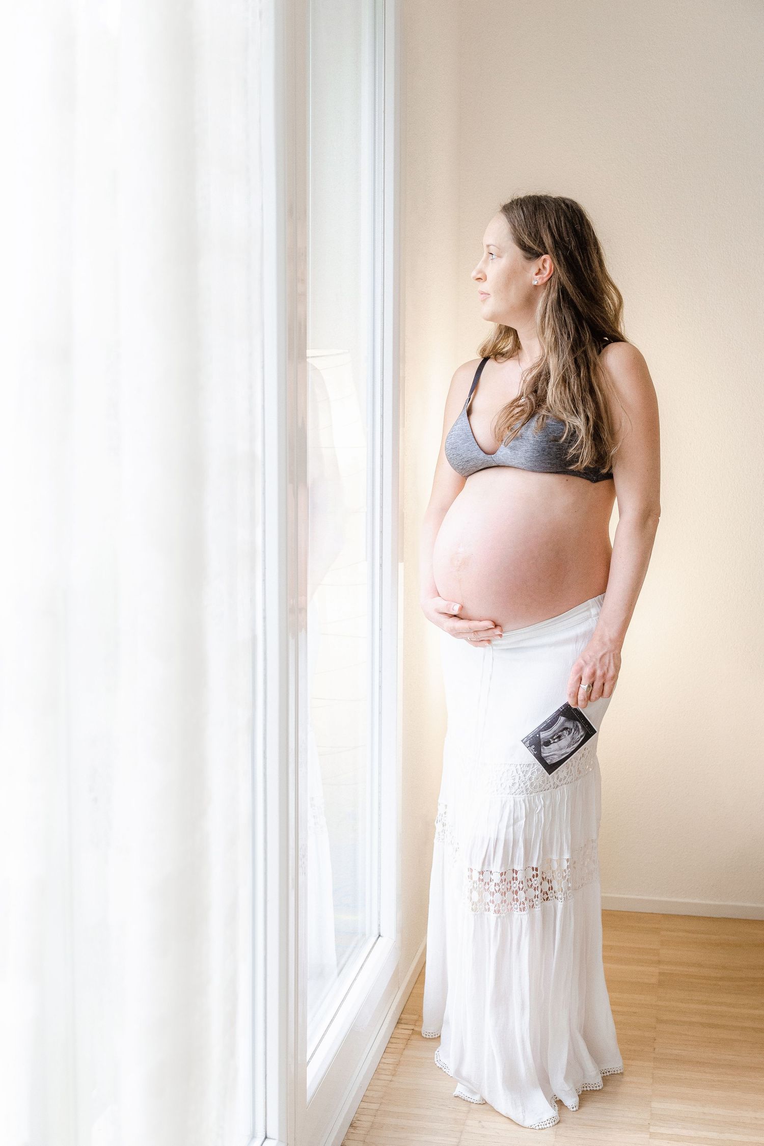Expectant mother in grey bralette and flowing white maxi skirt stands by window holding ultrasound photo in natural light.