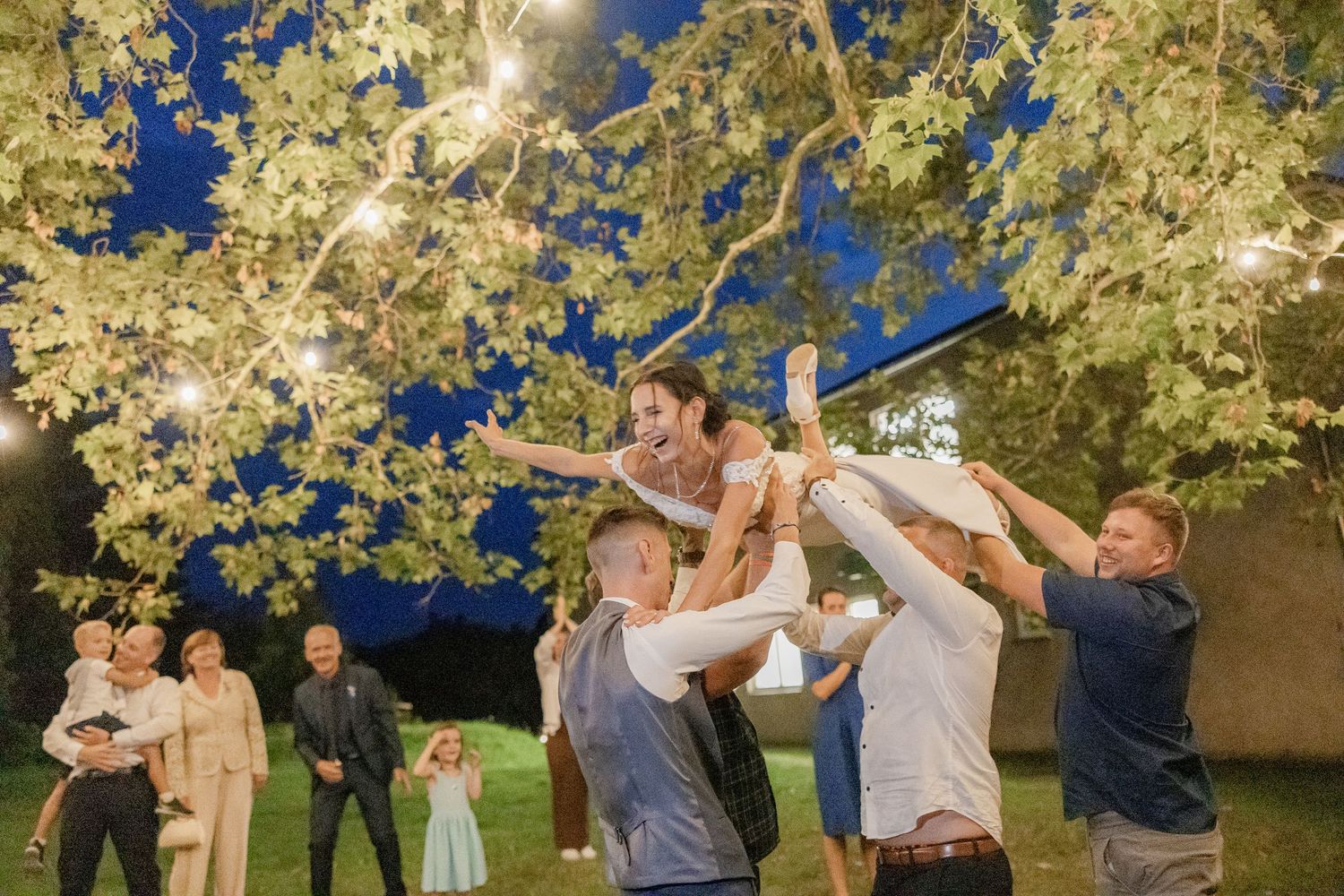 A group of people in light colored formal attire celebrate joyfully under illuminated trees at dusk.